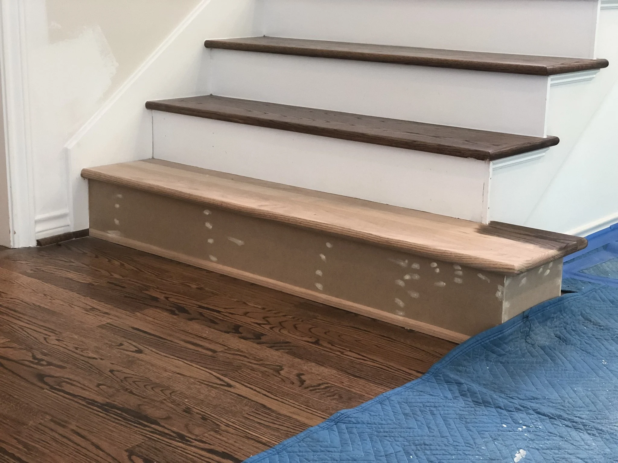 Wooden staircase under renovation with a partially sanded step and unfinished riser on hardwood flooring, covered partially with a protective blue mat.