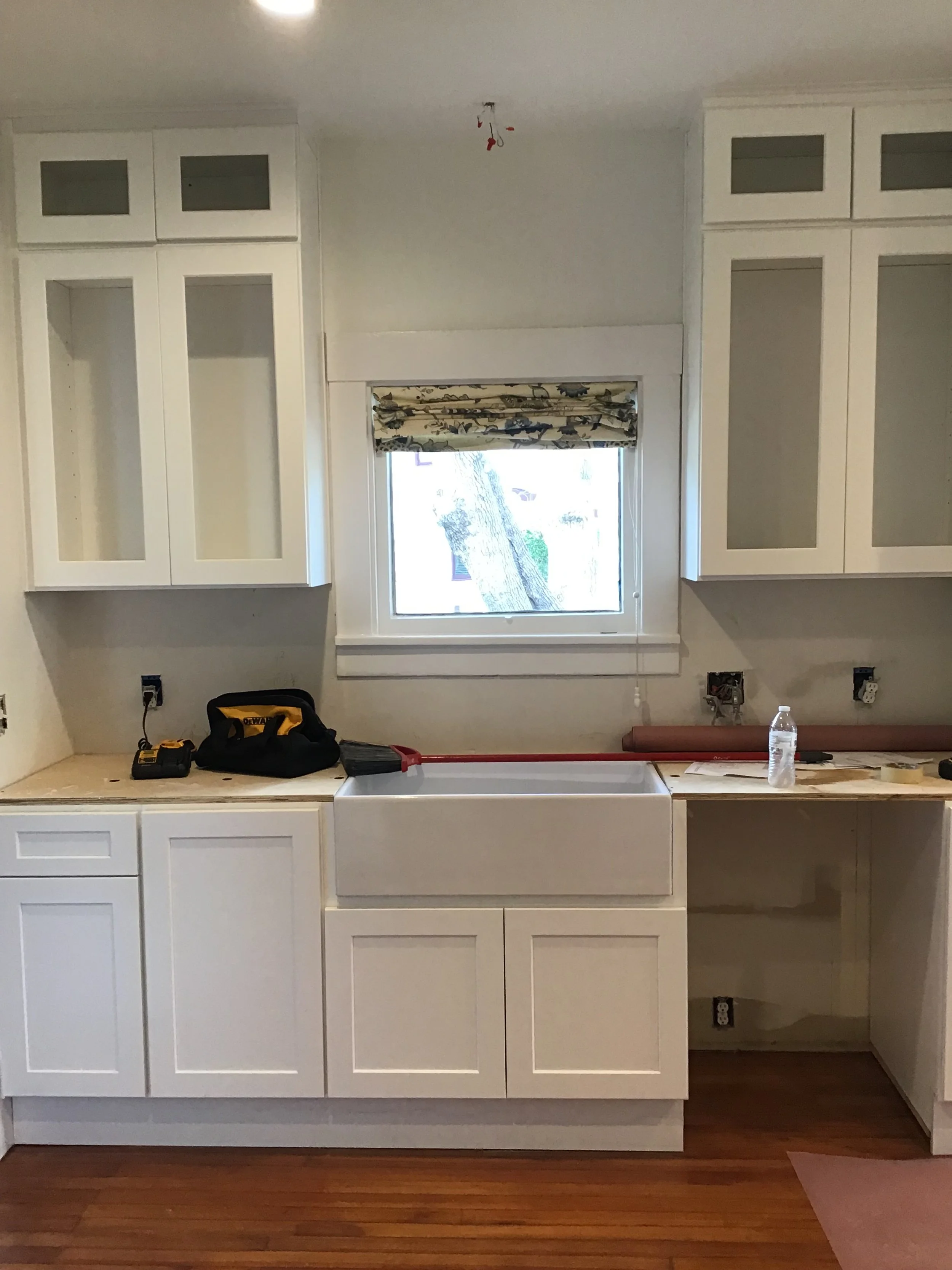 Kitchen under renovation with white cabinets, a large farmhouse sink, a window with a patterned valance, and remodeling tools on the countertop.