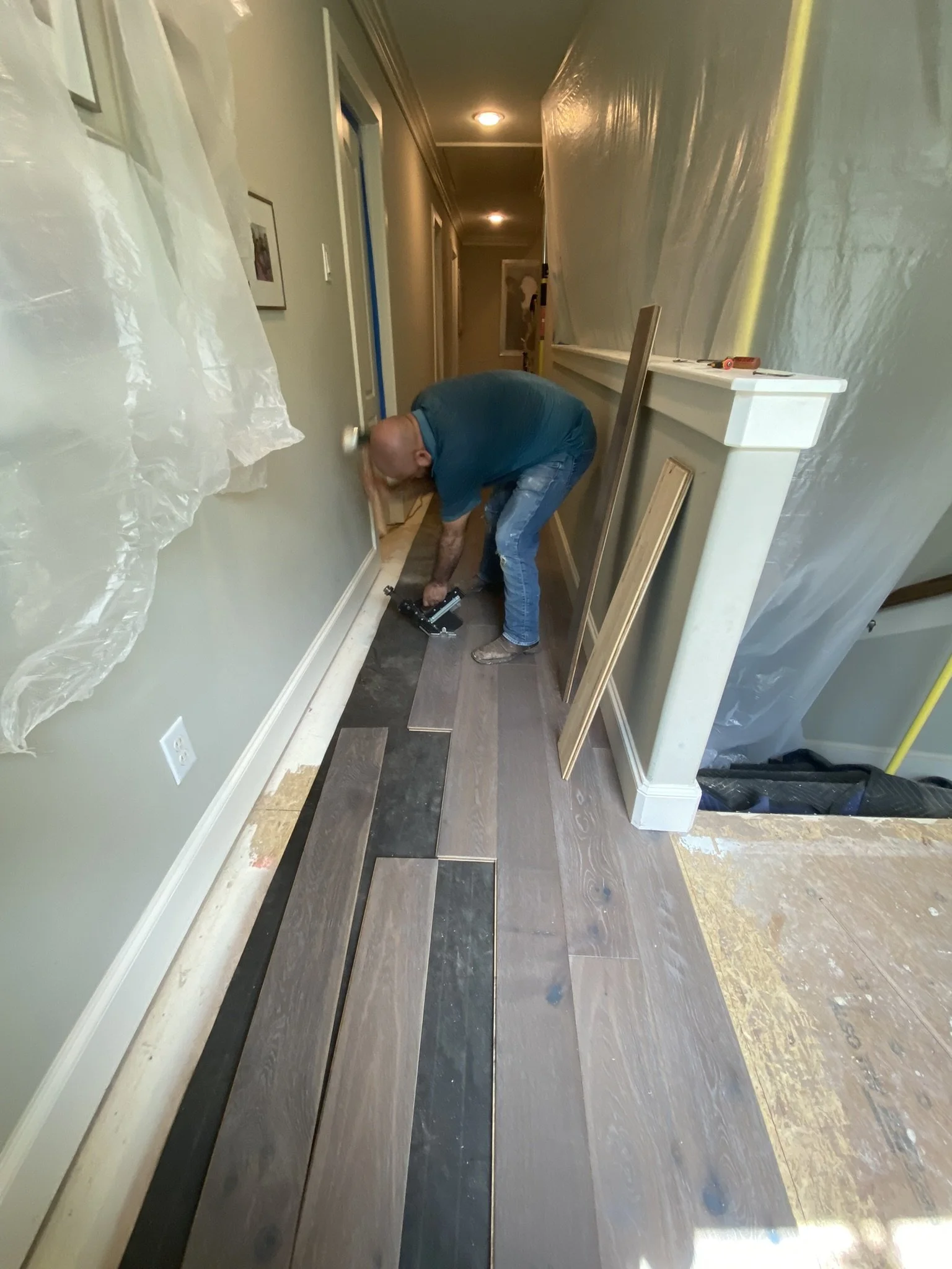 A person installing wooden flooring in a hallway. They are using a tool for the installation. The area has plastic coverings on one side, and some planks and tools are visible on the floor.