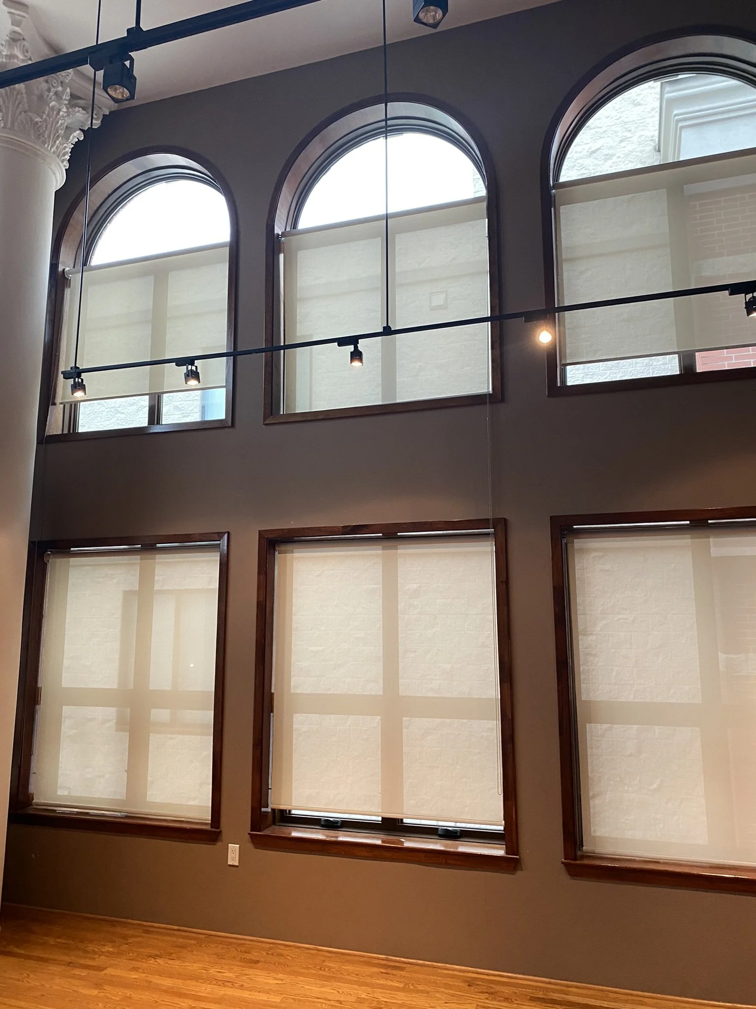 Interior view of a room with brown wall, showcasing large arched windows at the top and rectangular windows below, all with white shades. The room has a wooden floor, overhead track lighting, and a white decorative pillar on the left.