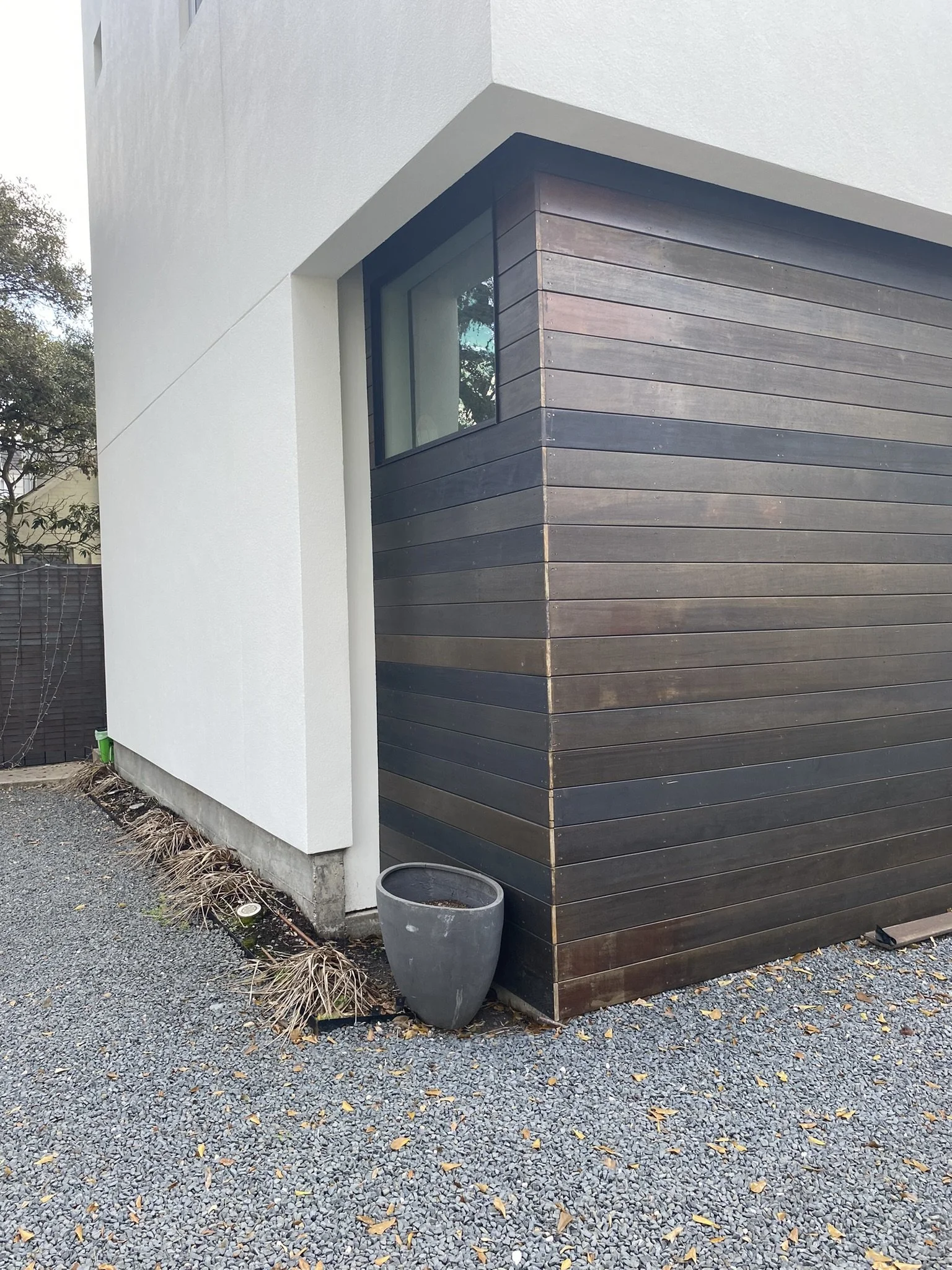 Modern house corner with wood siding, white stucco wall, window, gravel ground, large empty planter, and dry plants along the wall.