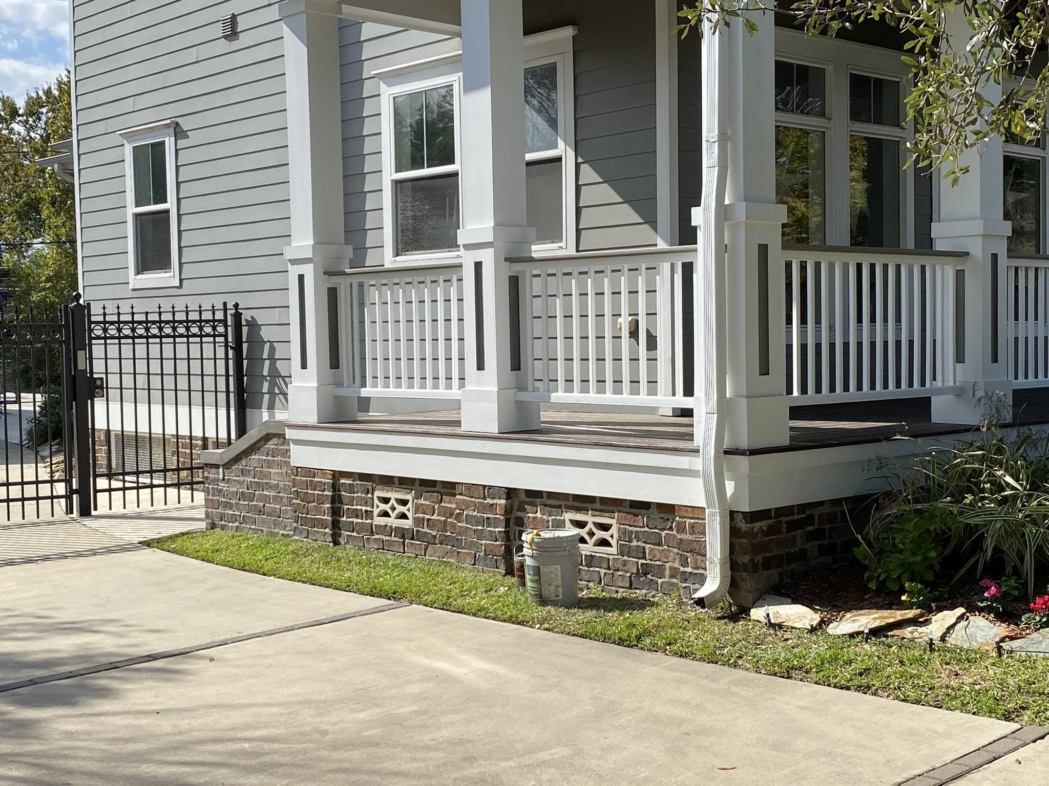 Exterior view of a house with a covered porch and white railing, gray siding, brick foundation, and a black metal gate beside a concrete driveway. The porch is decorated with some plants and flowers.
