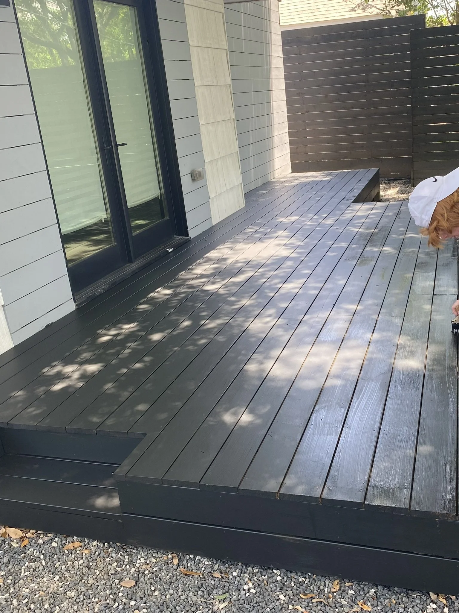 Outdoor wooden deck with dark wood finish, adjacent to a house with light siding and glass door. Fenced area in the background, person partially visible on the right.