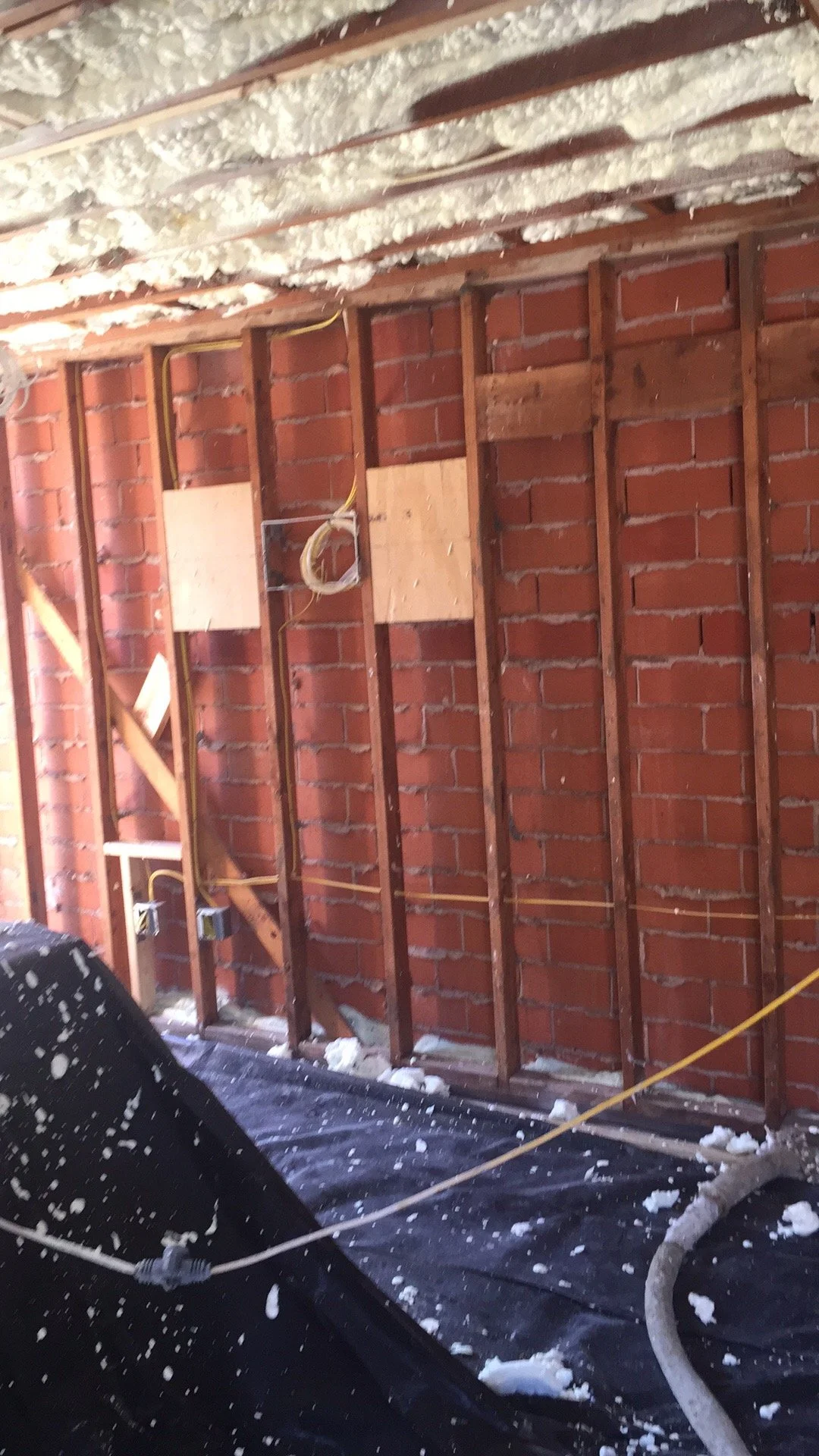 Interior of a room under renovation with exposed brick walls, wooden studs, and spray foam insulation on the ceiling.
