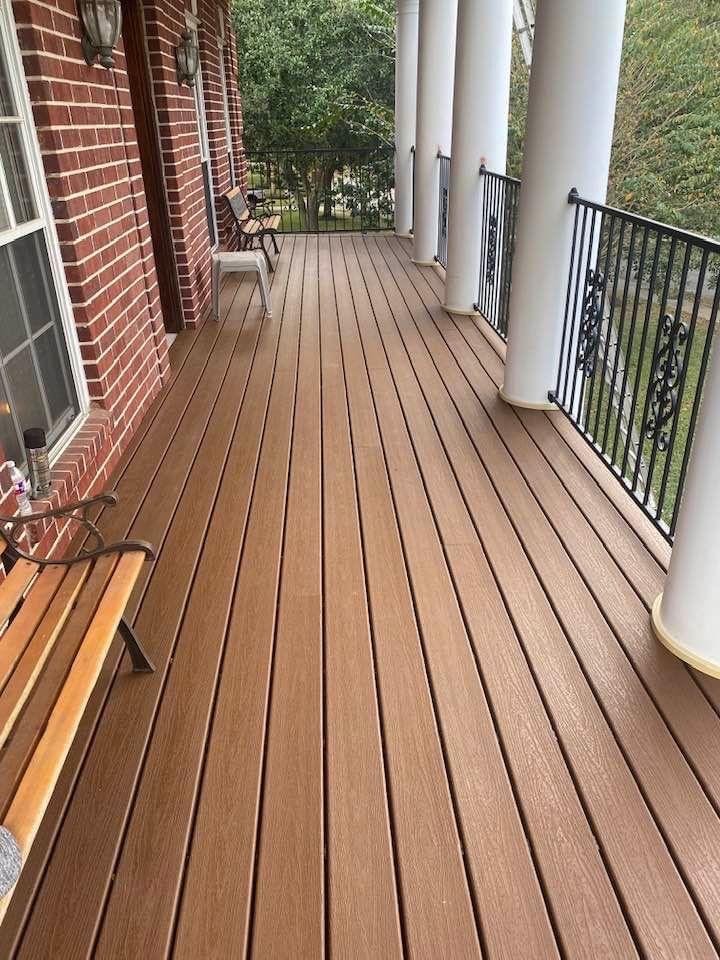 Wooden porch with white columns, wall lamps, a bench, and a chair against a brick wall, overlooking a garden with trees.