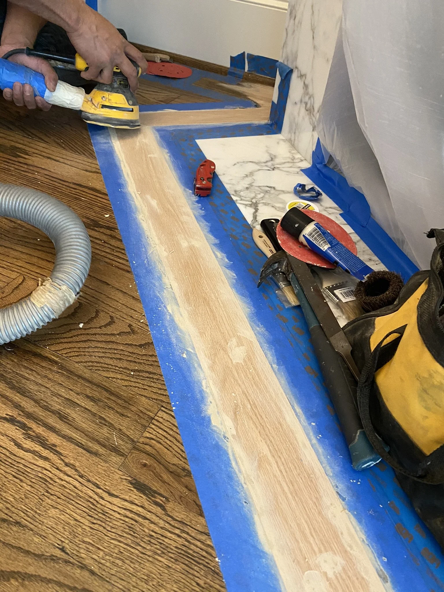 A person using an orbital sander on a wood floor with blue painter's tape around the edges, surrounded by various renovation tools including a hammer, chisel, utility knife, sanding discs, and adhesive tube. A dust collection hose is visible.