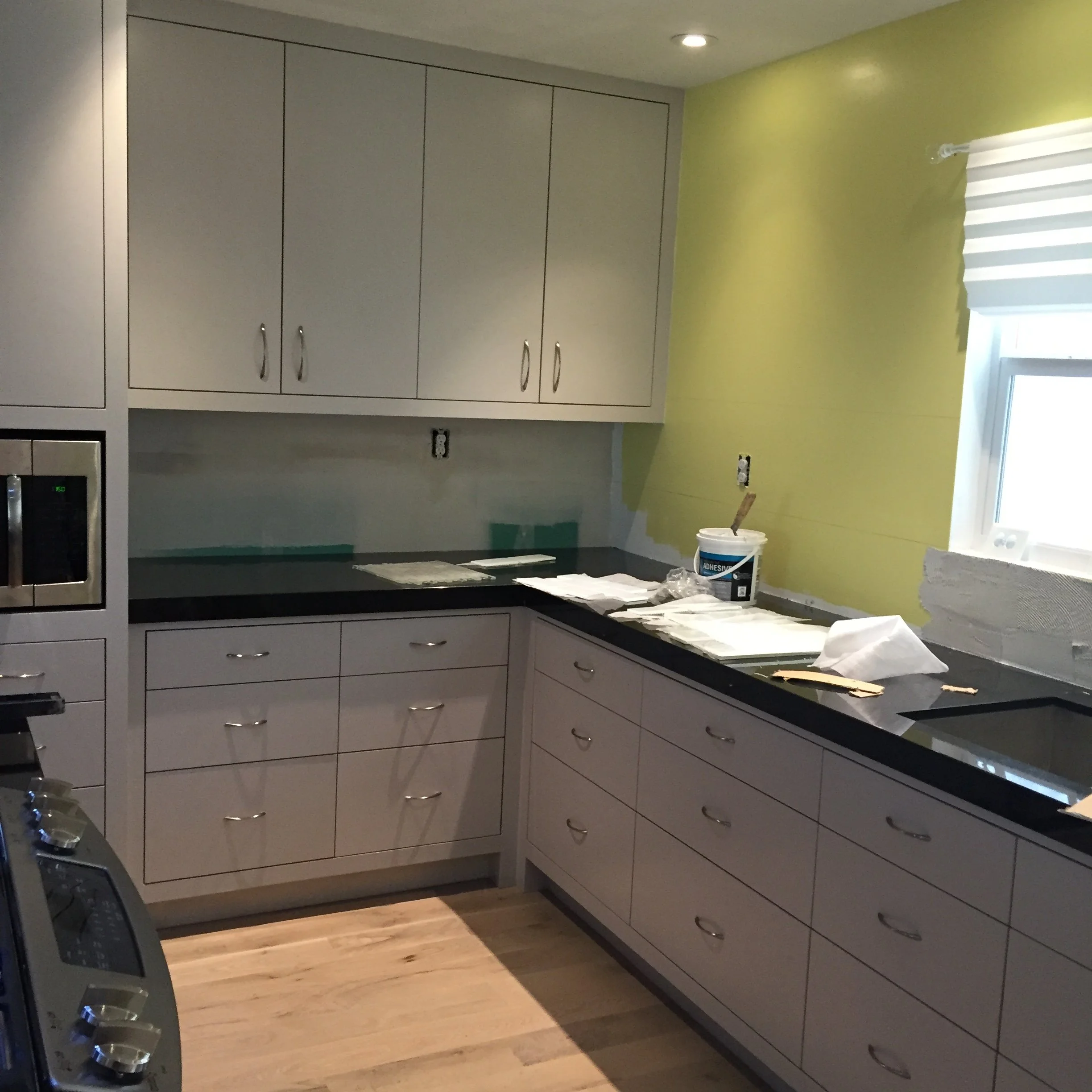 Kitchen interior with unfinished countertops, light gray cabinets, lime green wall, and a window with white blinds. A can of adhesive and trowels are on the counter, suggesting ongoing renovations.