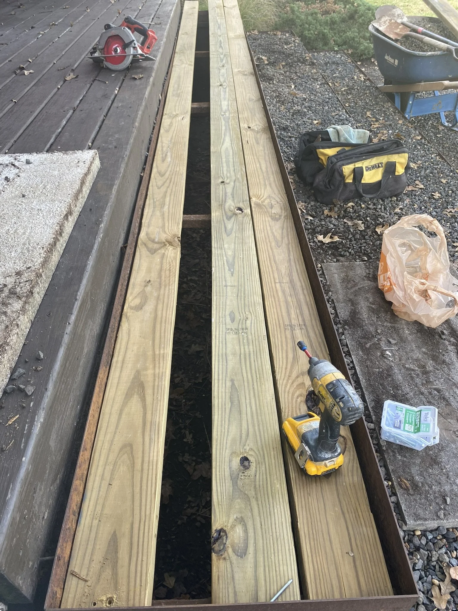 Deck construction scene with wooden planks, a power drill, a circular saw, and a tool bag on a gravel surface.