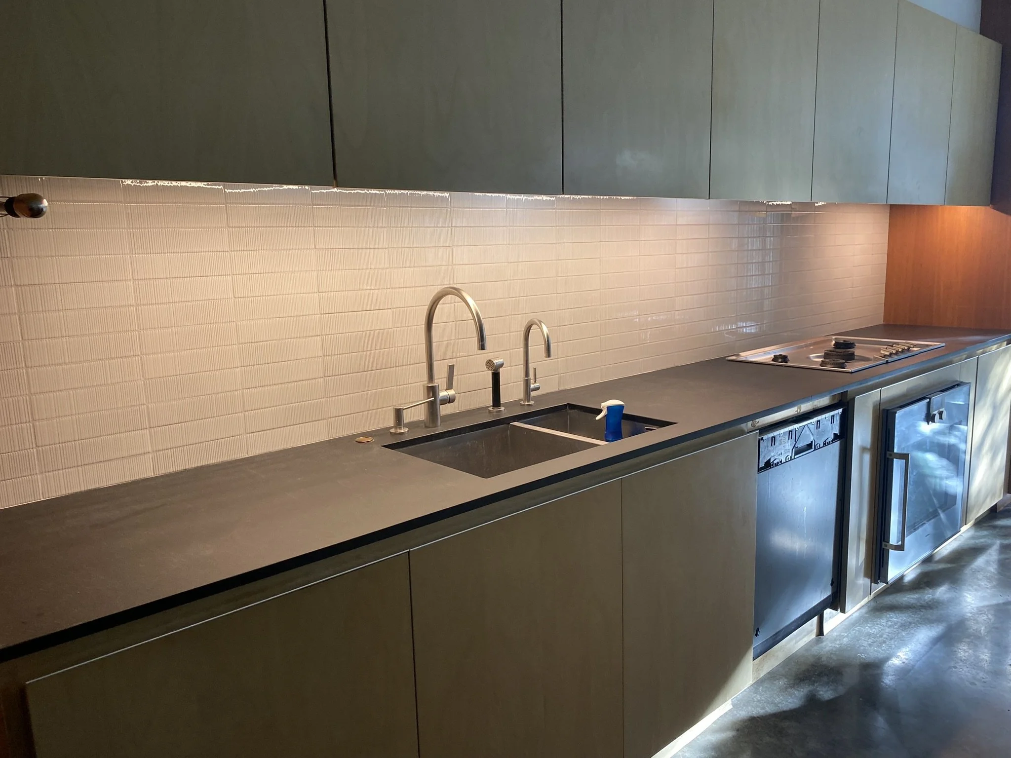 Modern kitchen with black countertop, stainless steel sink and faucet, under-cabinet lighting, closed dishwasher, oven, and light wood cabinets.