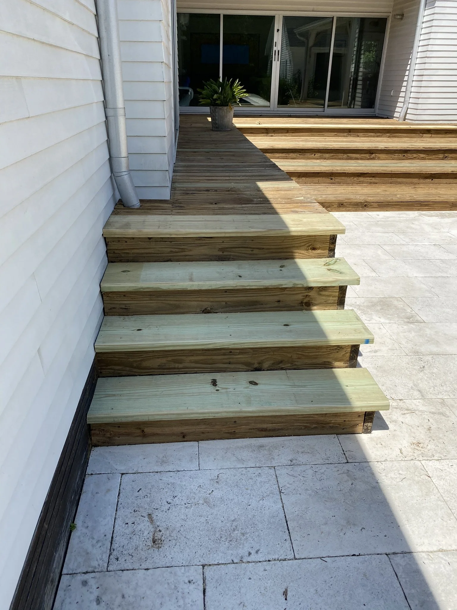 Wooden outdoor steps leading to a deck with sliding glass doors, white siding, and a potted plant on the deck.