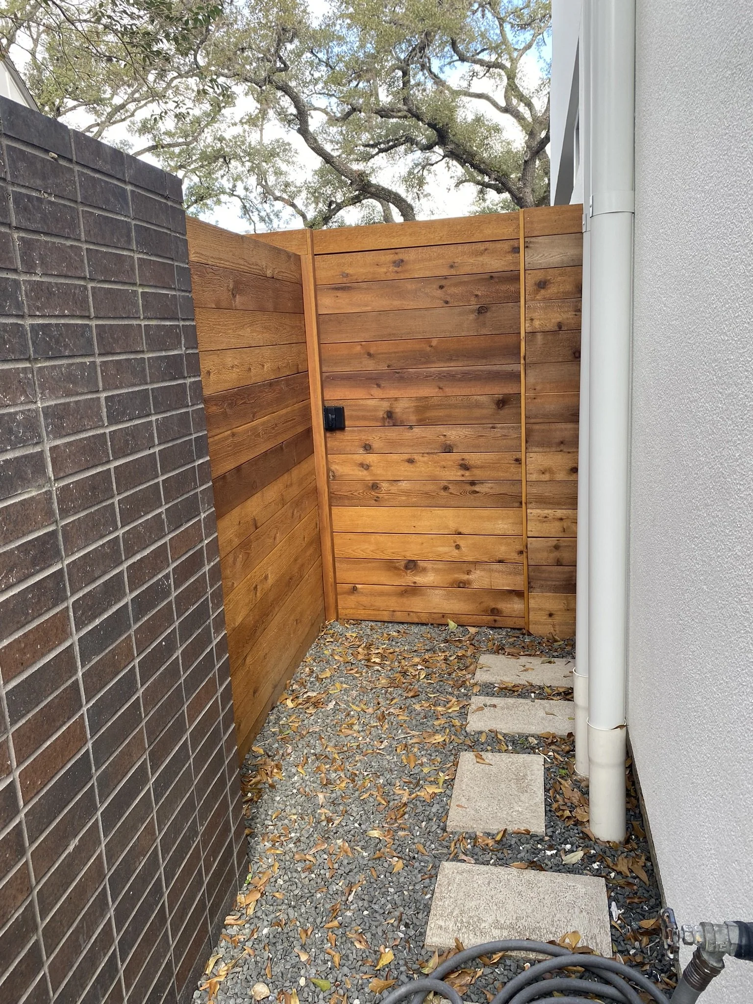 Outdoor fenced area with wooden privacy fence, gravel ground, fallen leaves, stepping stones, and garden hose.