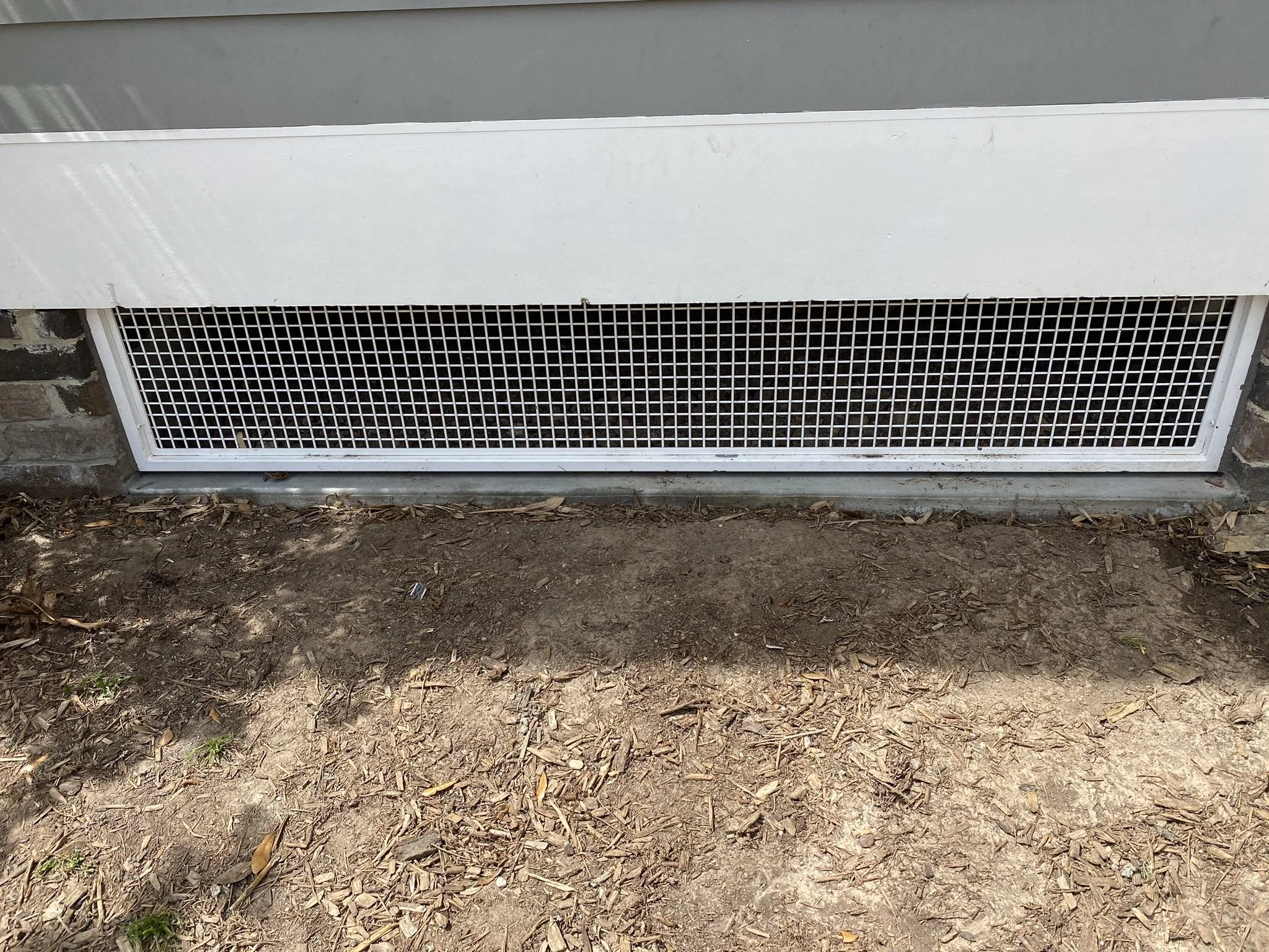 Crawl space vent with white metal grid on a house exterior near soil and mulch.