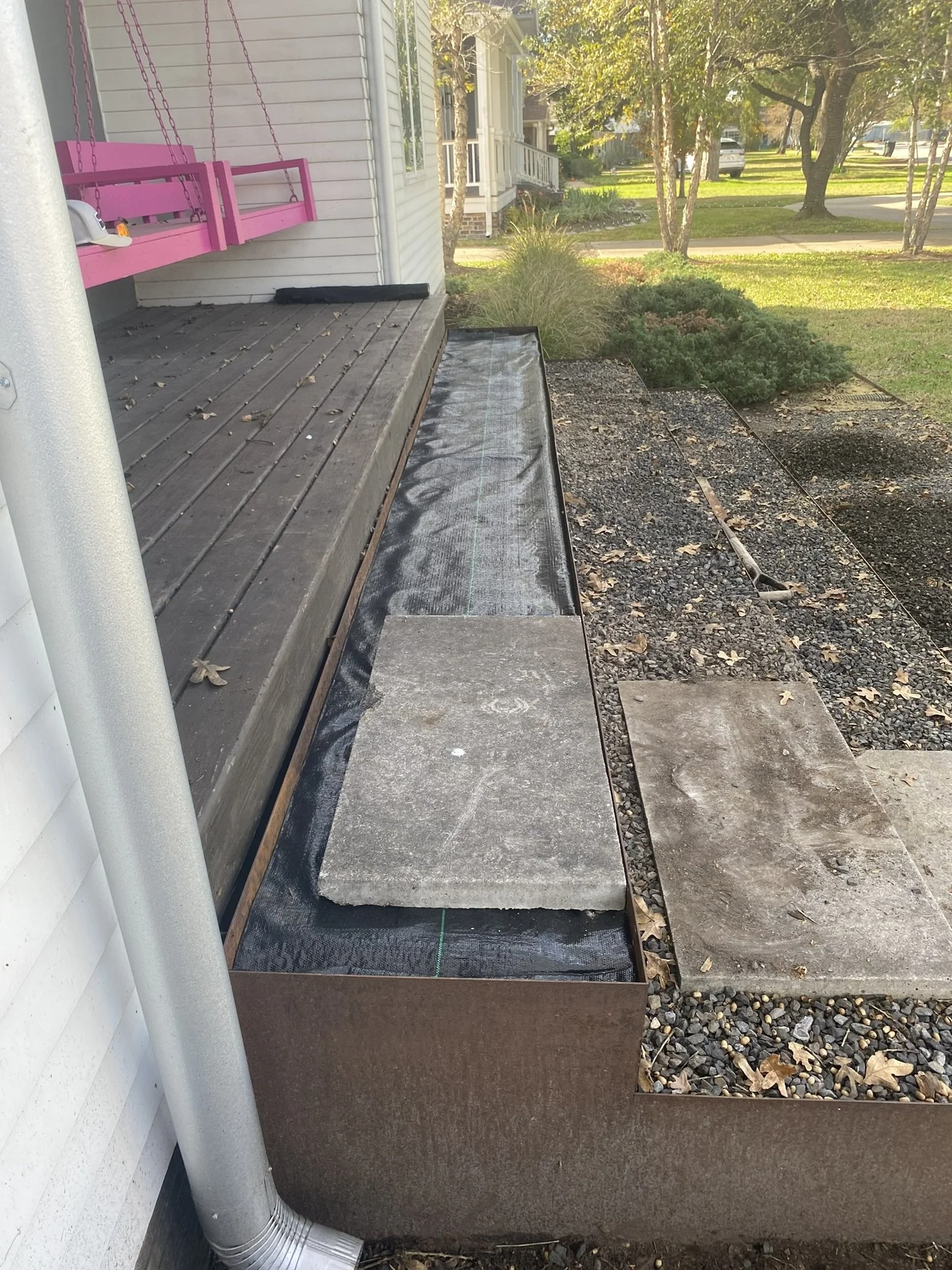 Front porch with a pink porch swing and landscaping under construction, featuring concrete slabs, gravel, and a black weed barrier. A silver downspout is situated on the left.
