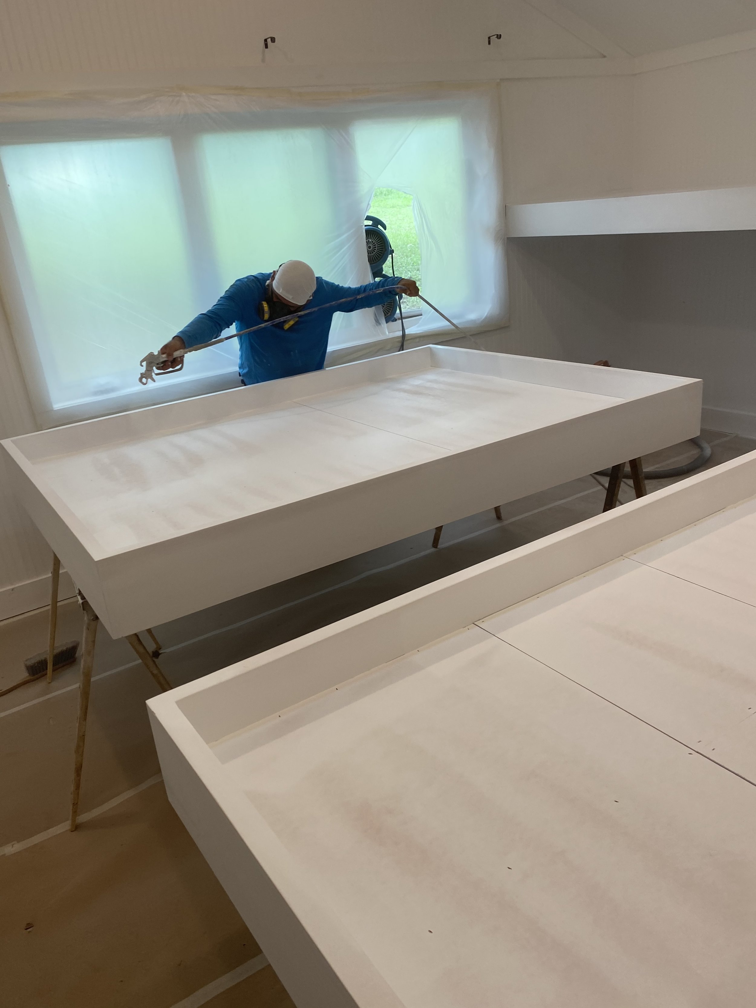 Person in protective gear spray painting a large white surface in a room with windows covered in plastic.
