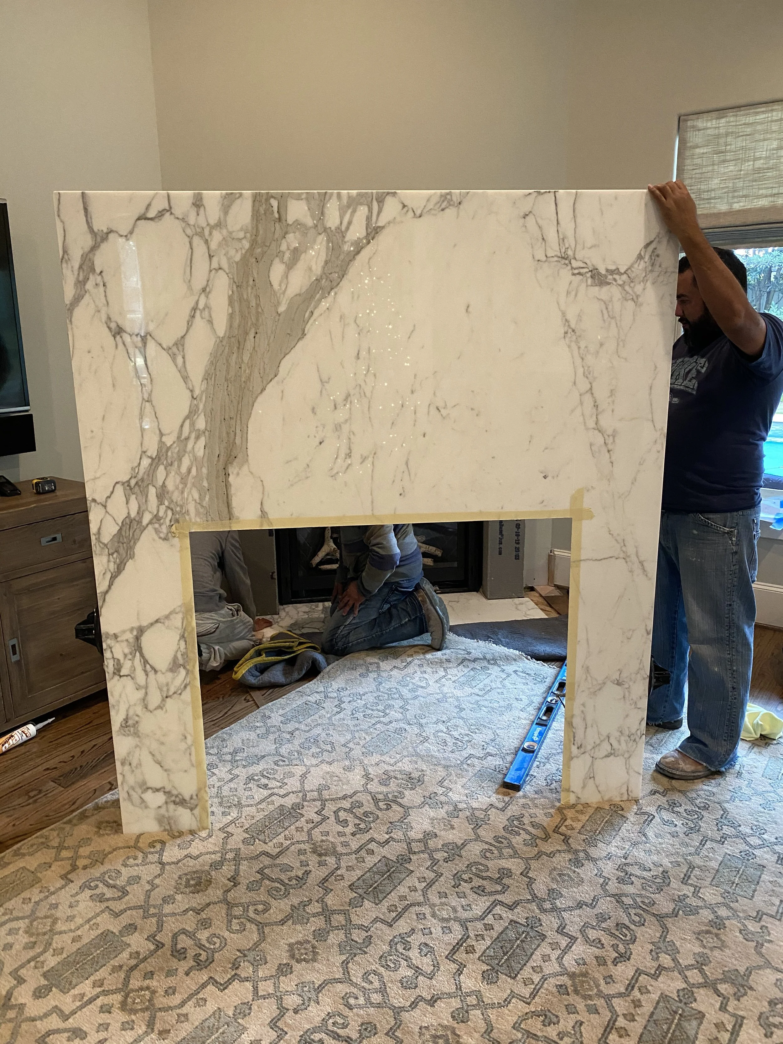 Installation of a white marble fireplace facade with gray veining, supported by two people in a living room. A patterned rug is on the wooden floor, and tools are scattered around.