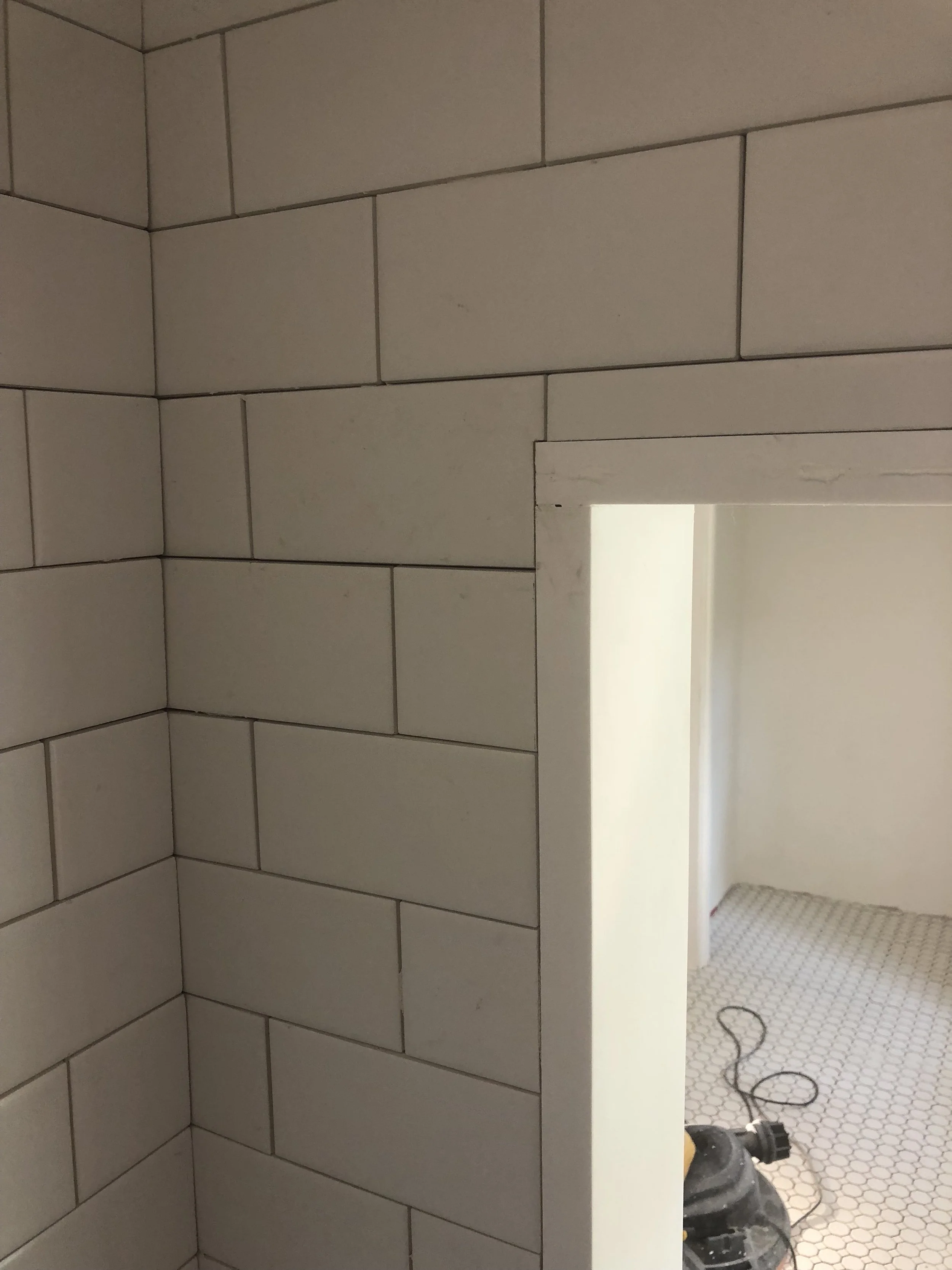 Corner of a tiled wall with white rectangular tiles, adjacent to a doorway leading to a room with hexagonal floor tiles.