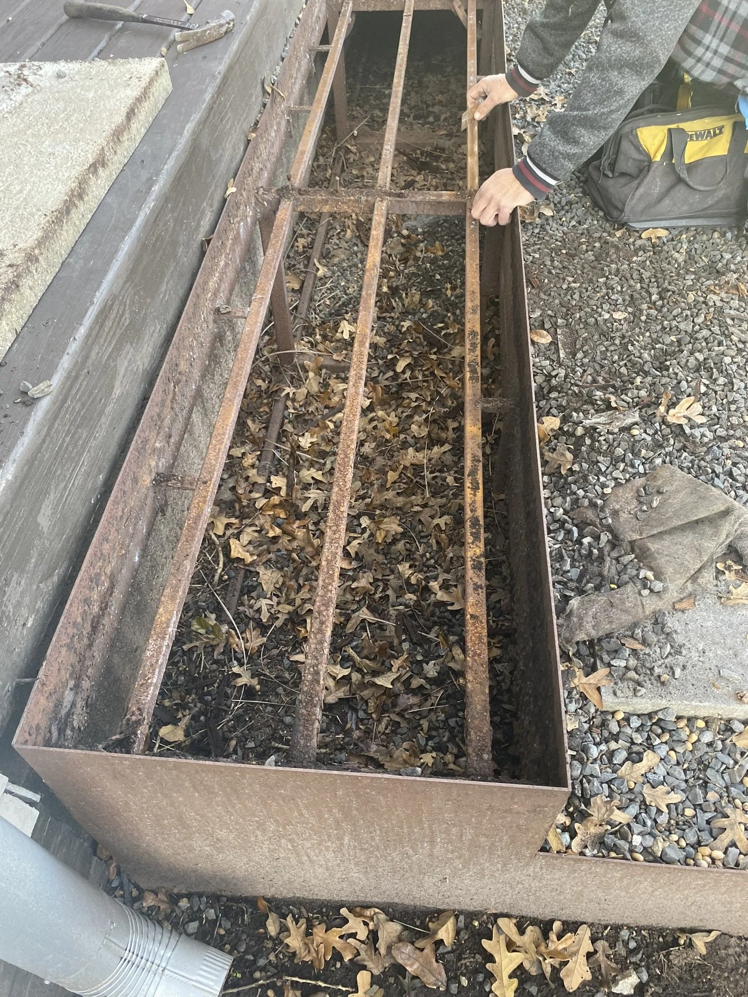 A person working on a rusty metal frame filled with dried leaves and debris, near a wooden deck. Tools and a tool bag are nearby on gravel.