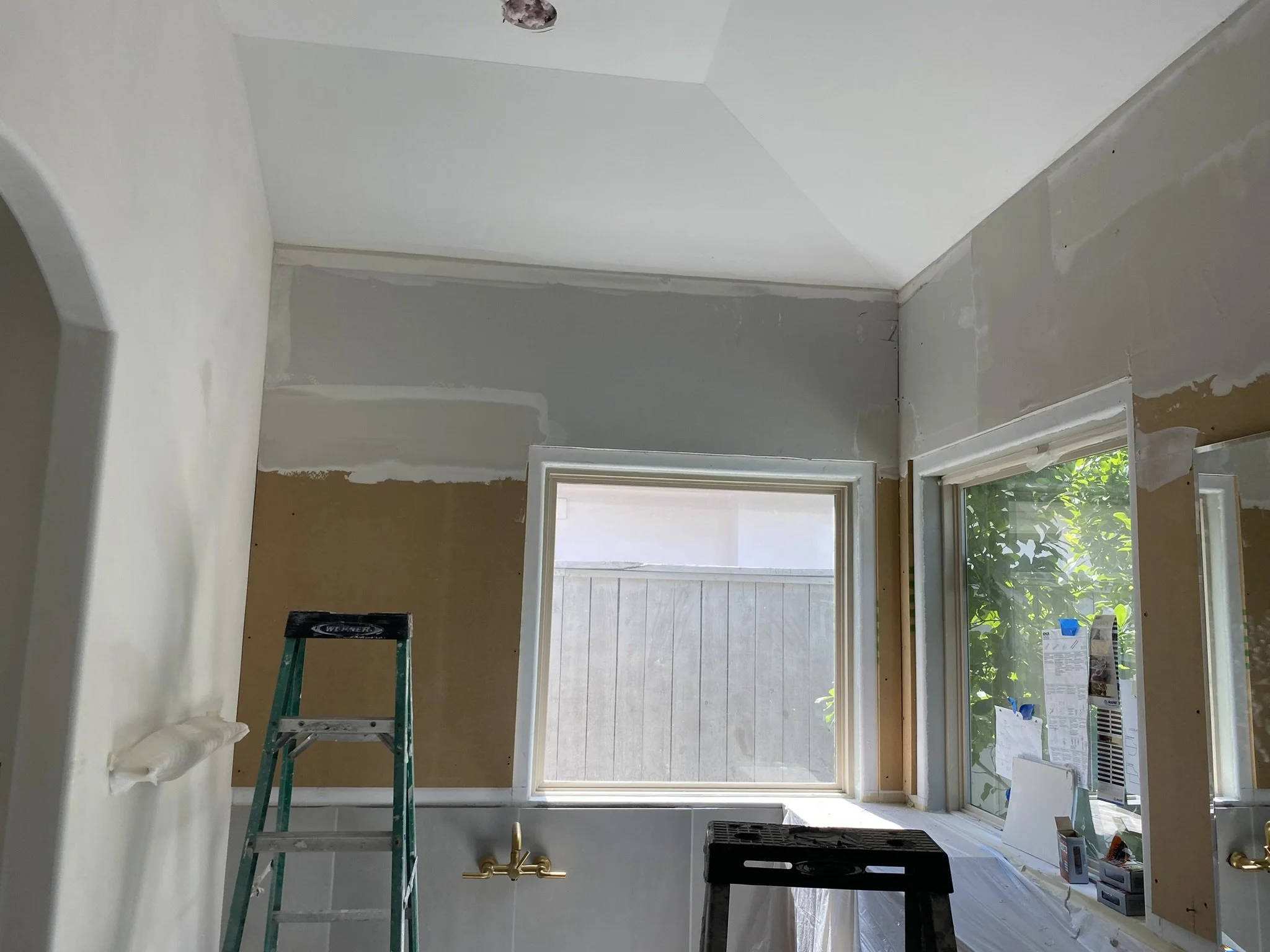A partially renovated room with unfinished walls, two windows, and a high ceiling. A ladder and a step stool are present, and the lower part of the walls is covered with protective material. Tools and renovation materials are visible near the window.