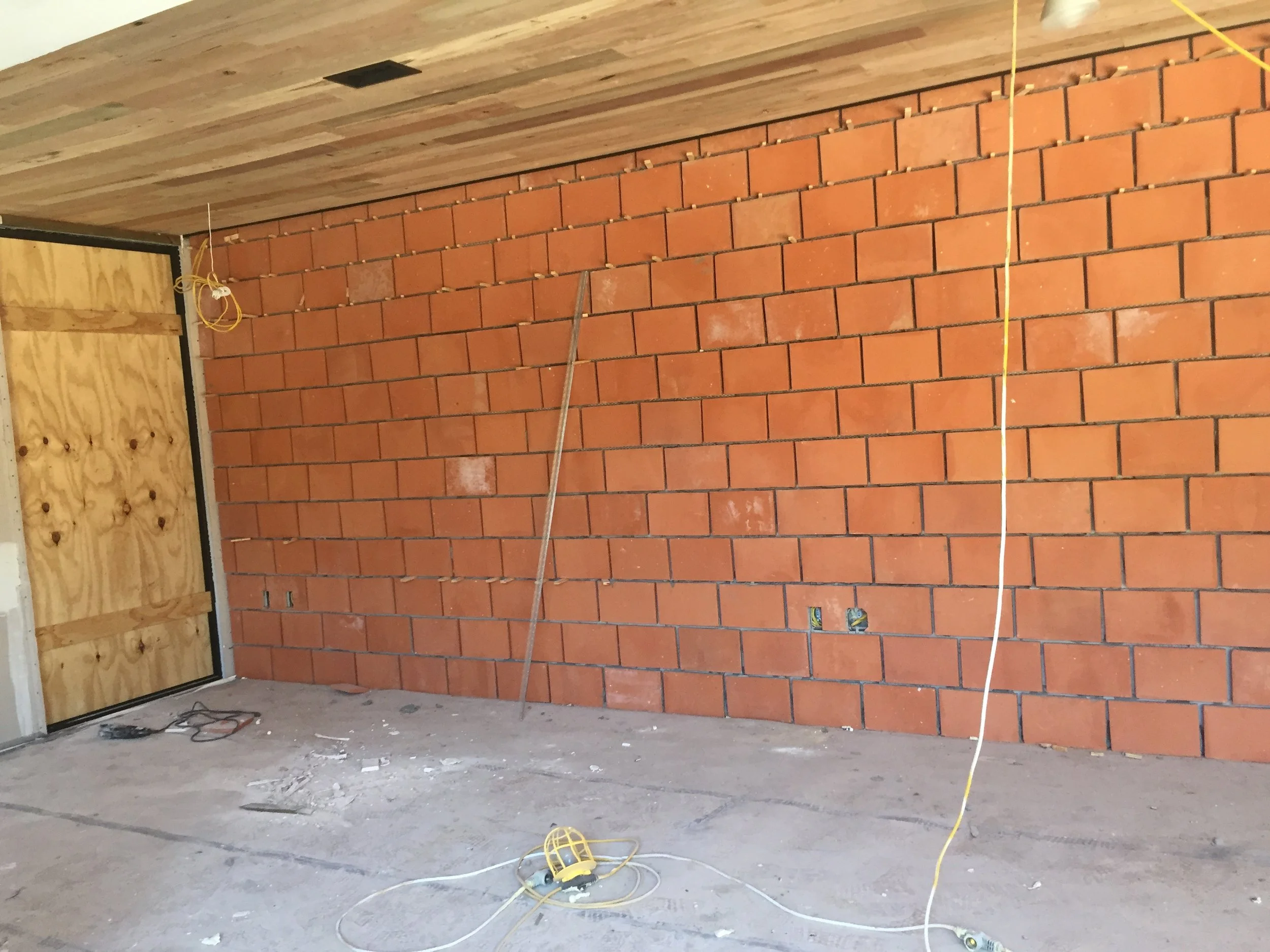 Partially constructed interior with unfinished brick wall, wooden ceiling, plywood-covered doorway, hanging electrical wires, and tools on the floor.