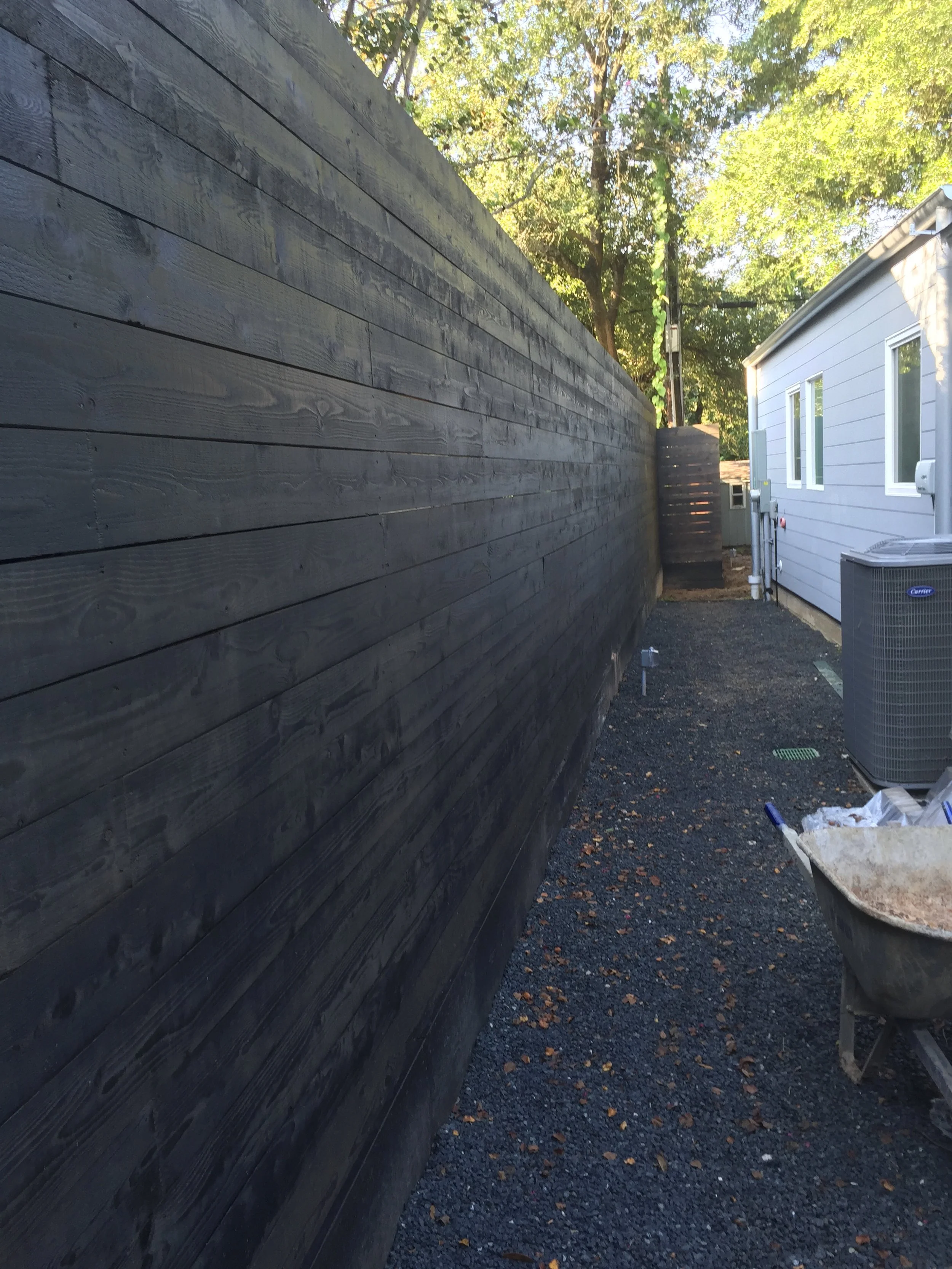 Narrow outdoor space with a tall black wooden fence on one side and a gray house wall on the other, gravel ground, trees in the background, and a wheelbarrow in the foreground.