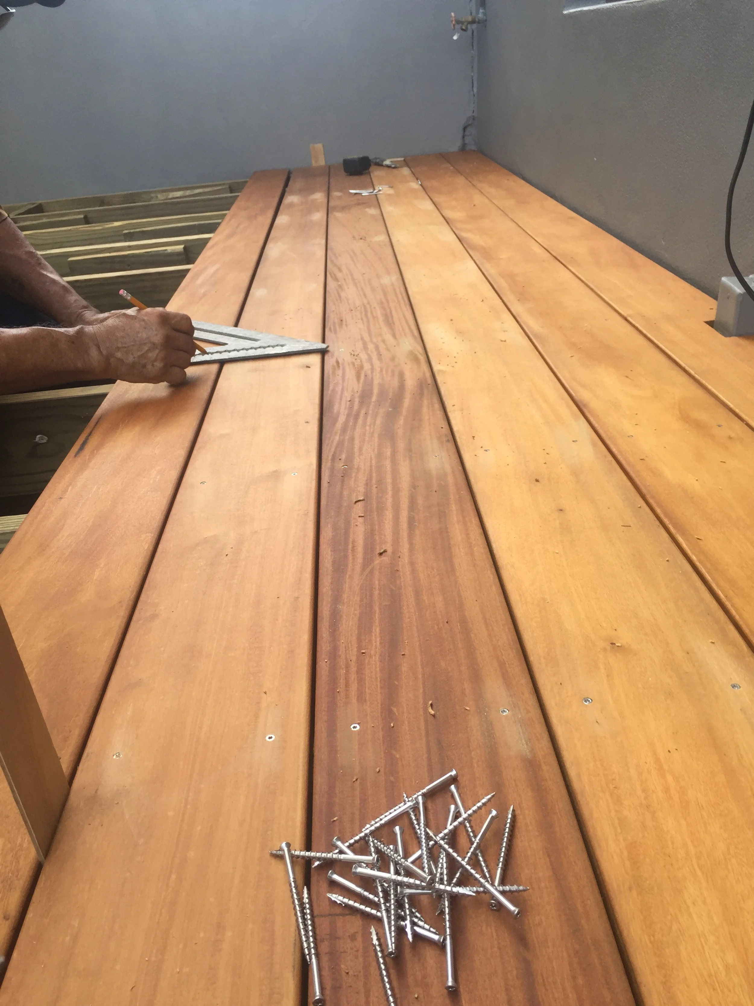 Person marking a wooden deck with a square tool and pencil, with screws and a rubber mallet nearby.