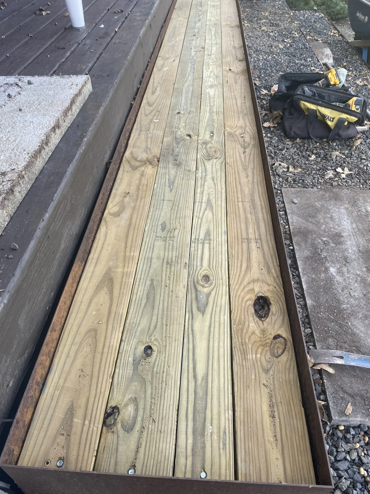 Wooden deck or pathway under construction with adjacent tool bag on gravel surface.
