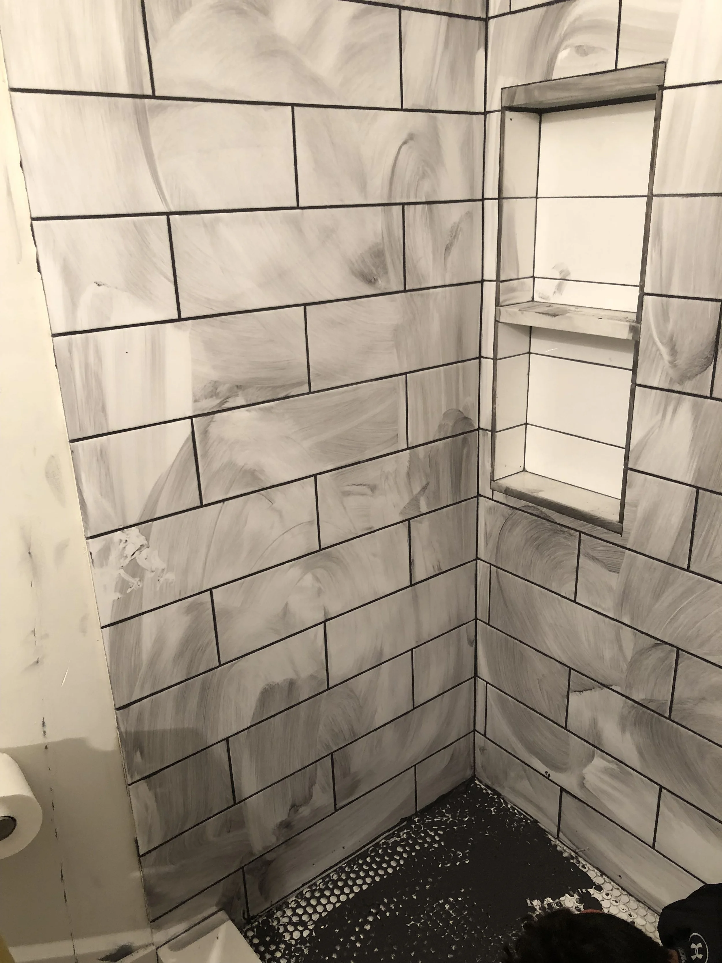 Shower with white subway tile walls and black grout, partially grouted with dark substance, and built-in shelf, under construction.
