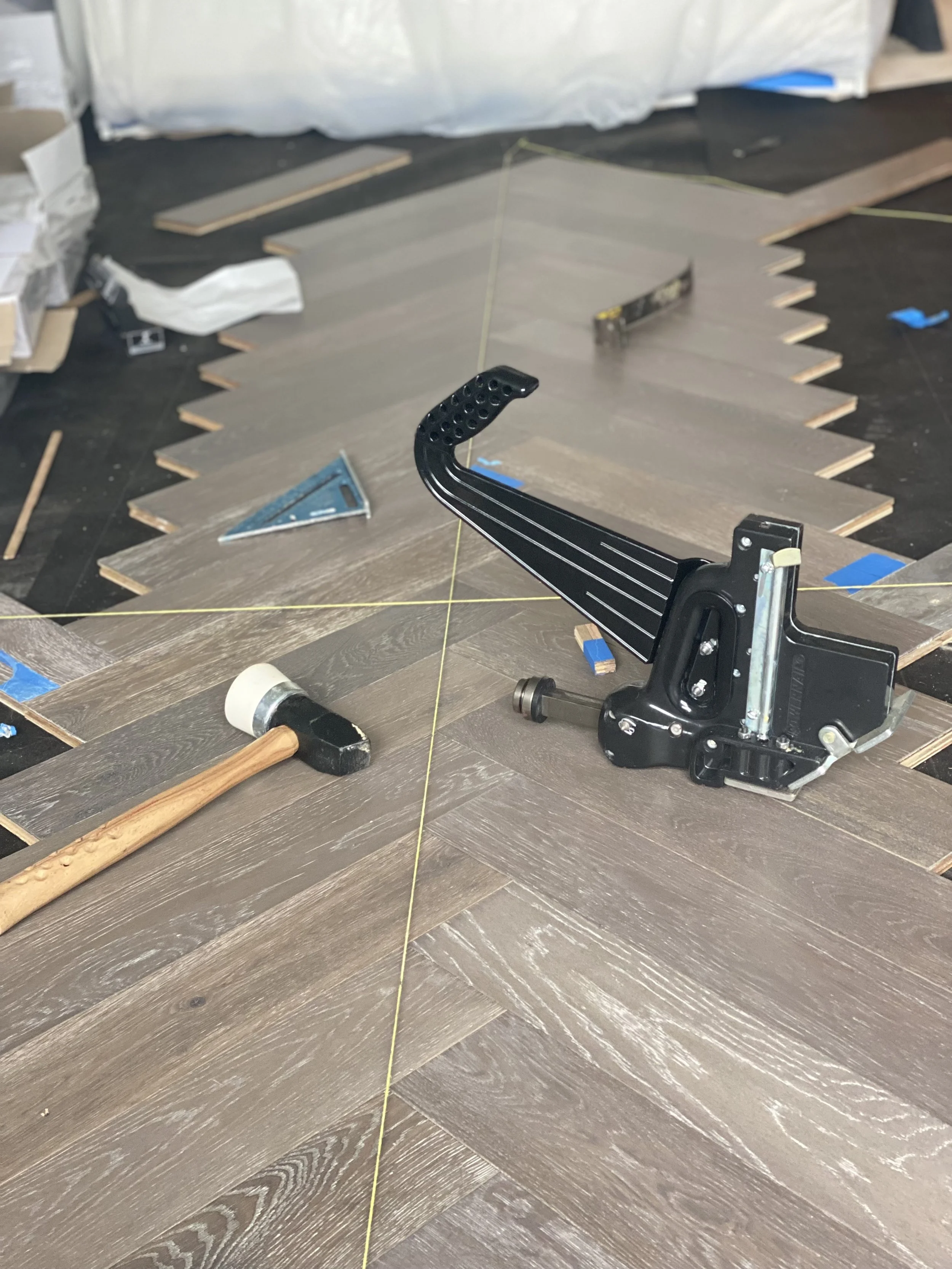 Floor installation scene showing herringbone wood pattern with a pneumatic flooring nailer, rubber mallet, triangular square, and measuring line. Boxes and scattered wood pieces are visible in the background.