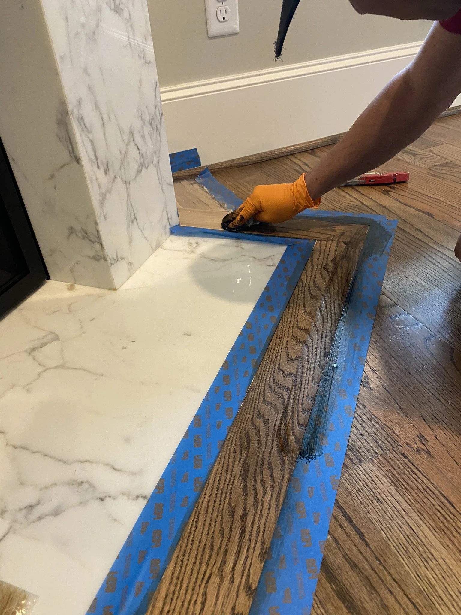 Person staining wood floor near a marble fireplace with blue painter's tape, wearing an orange glove.