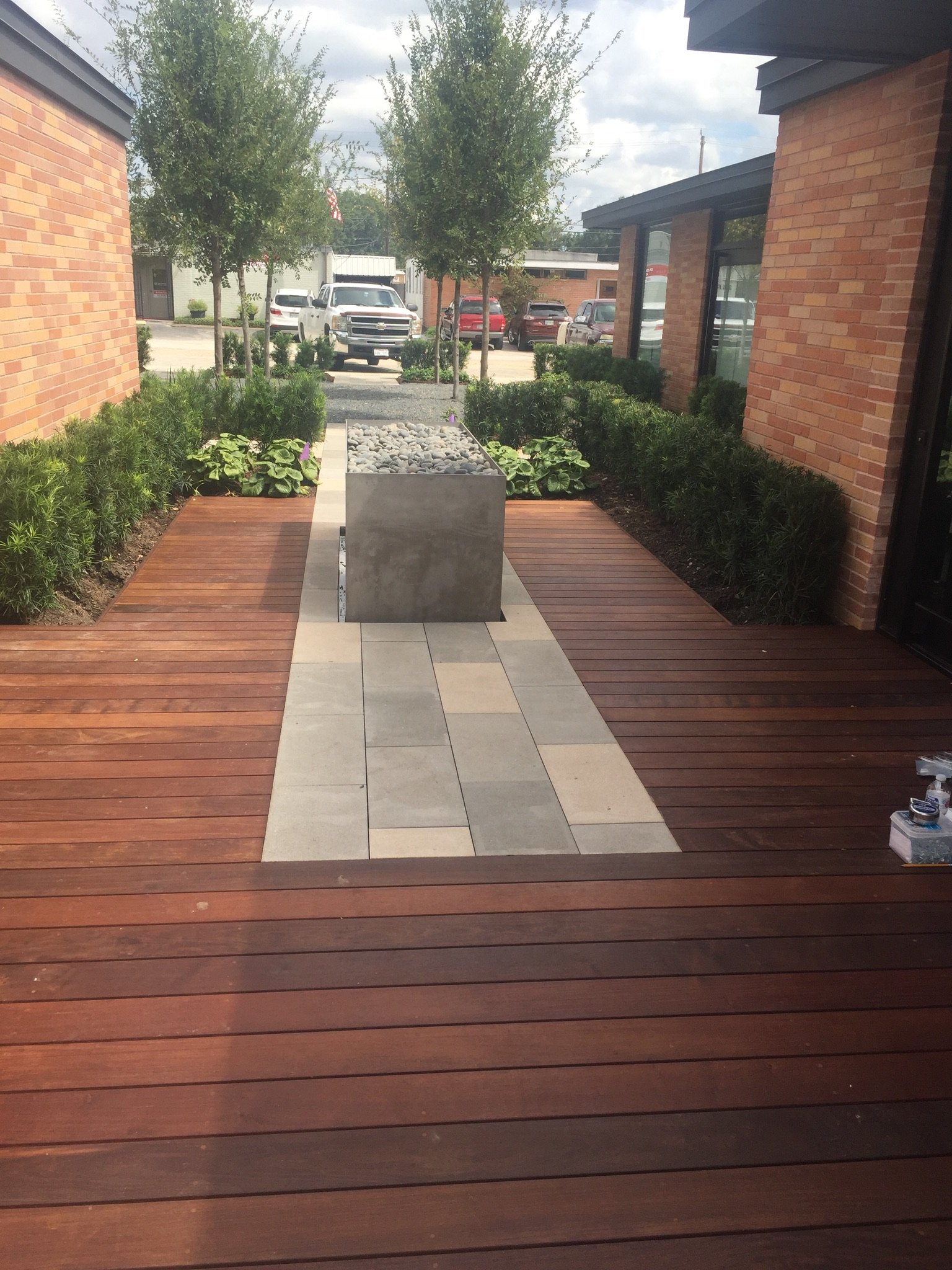 Modern outdoor walkway with wooden decking, stone pathway, and a centered cube planter filled with stones, surrounded by greenery and framed by brick walls.