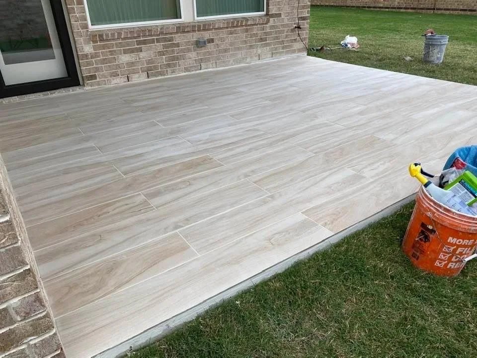 Outdoor patio with light wood-style tiles, adjacent to grass, a brick wall, and a bucket containing tools and supplies.