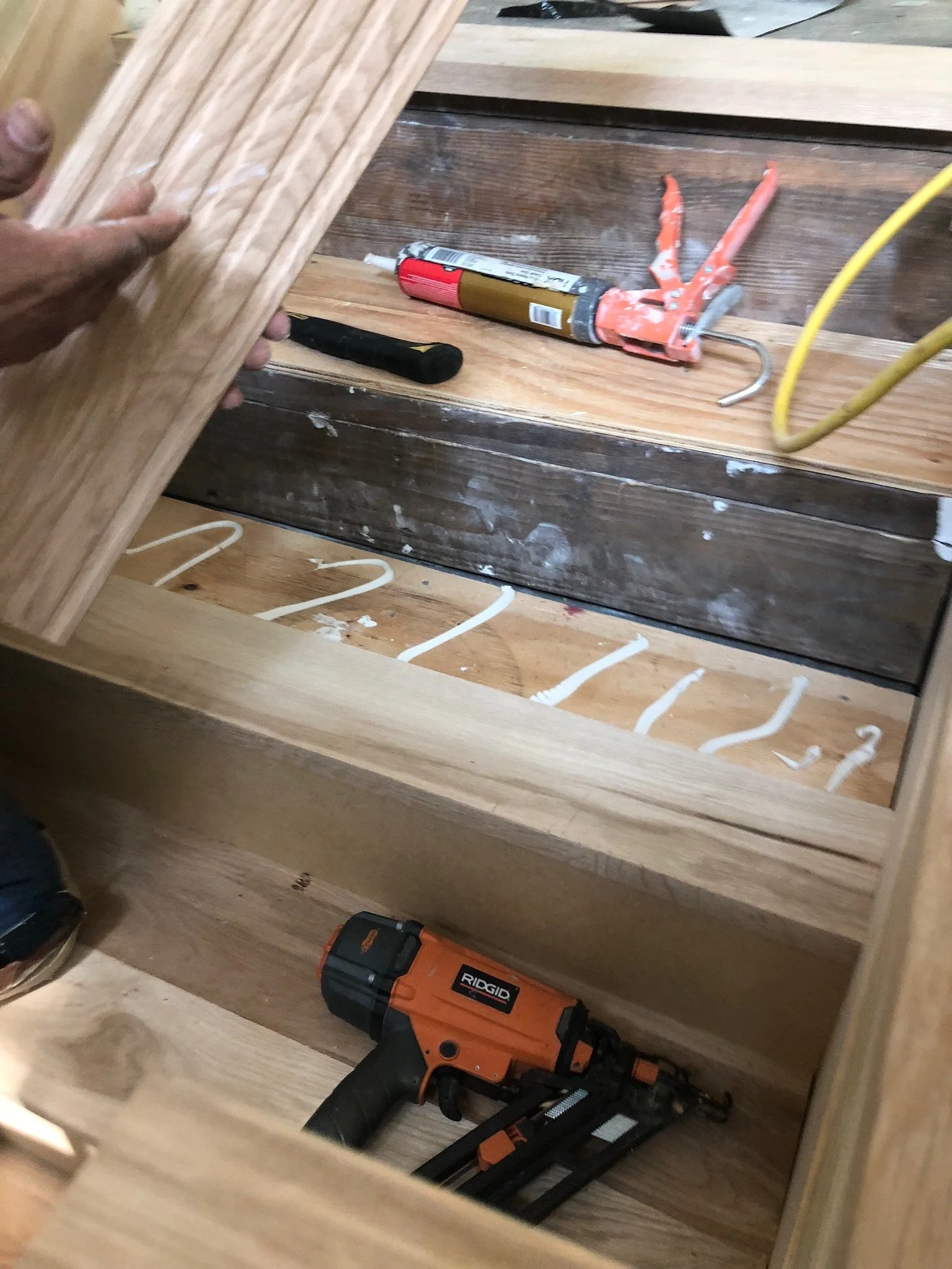 Close-up of staircase installation with tools and wood panel. A person is holding a wooden plank. A nail gun, caulk, and a hammer are visible.