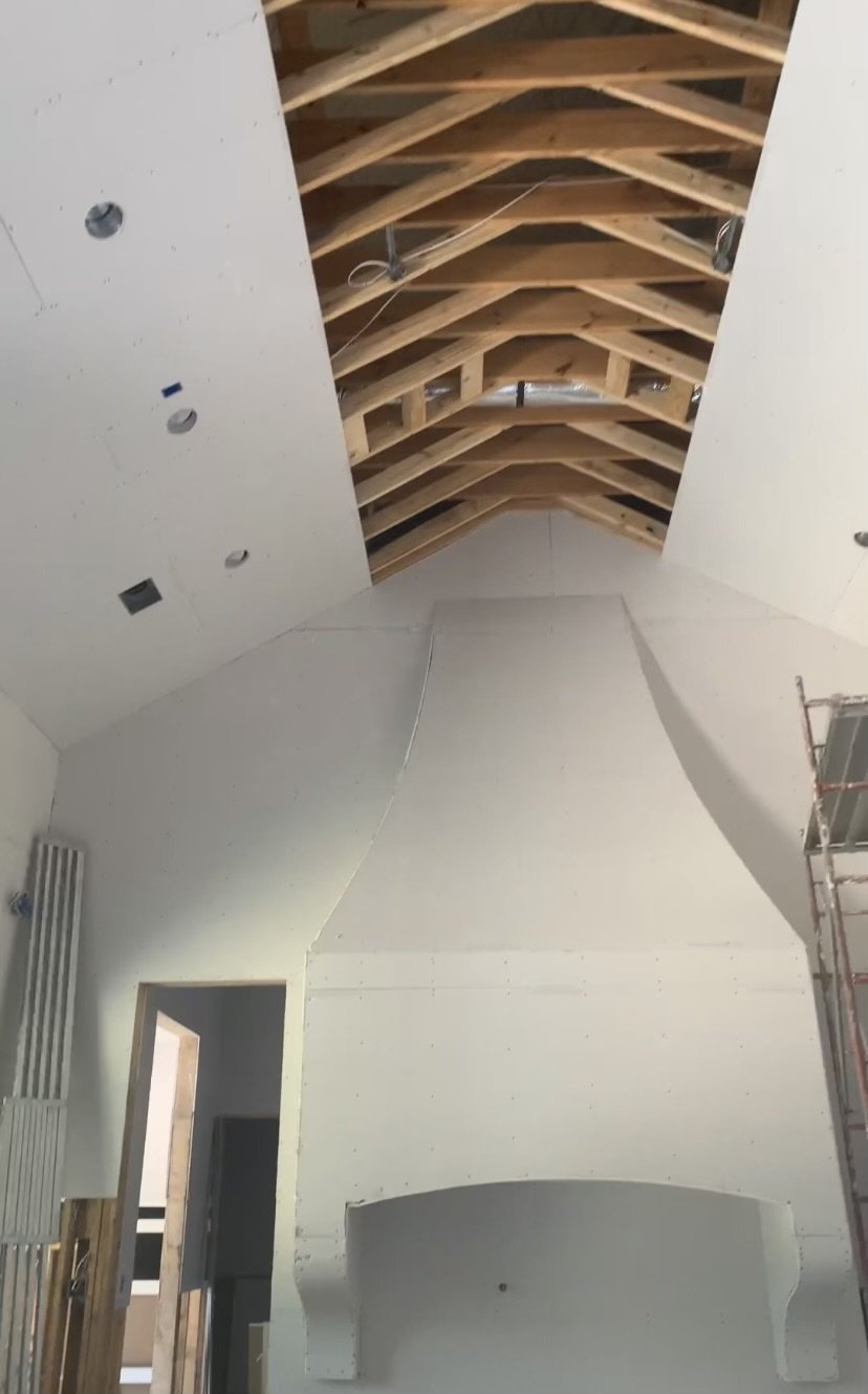 Interior construction view of a room with exposed wooden ceiling beams, drywall, and a large drywall structure appearing to be a fireplace or chimney. Scaffolding and unfinished construction materials are visible.