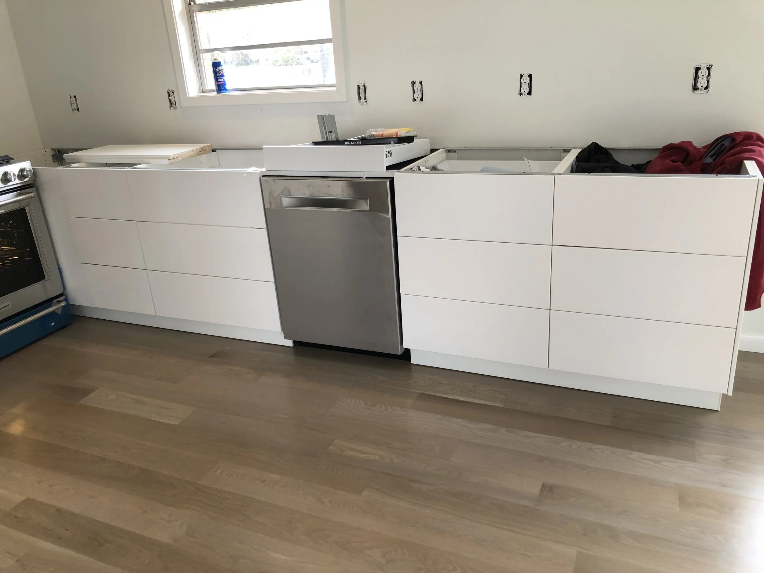 Modern kitchen with white cabinets, stainless steel dishwasher, and wood flooring. Unfinished wall sockets, stove, and construction materials visible.