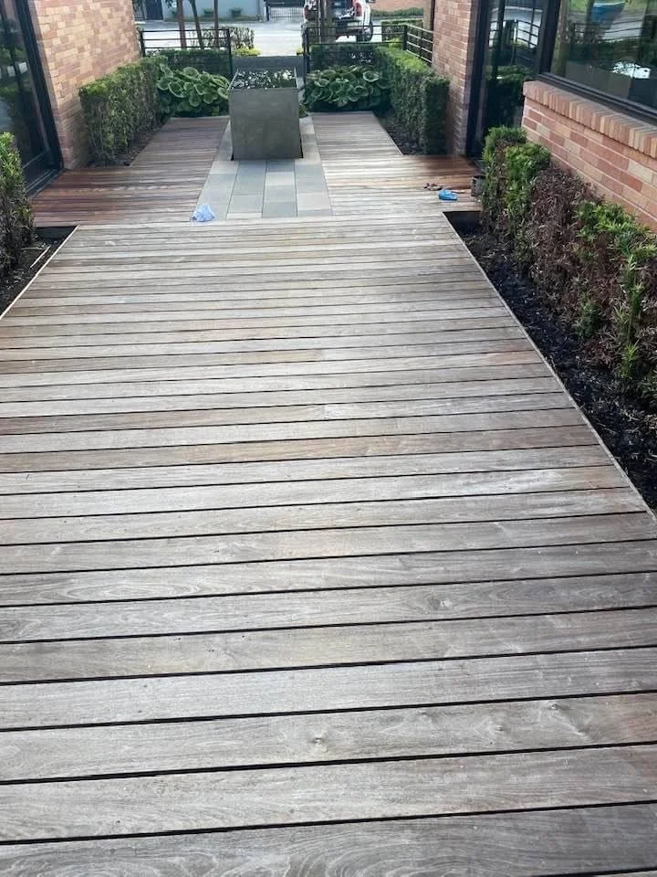 Wooden deck pathway with plants and a planter, surrounded by brick walls and windows.