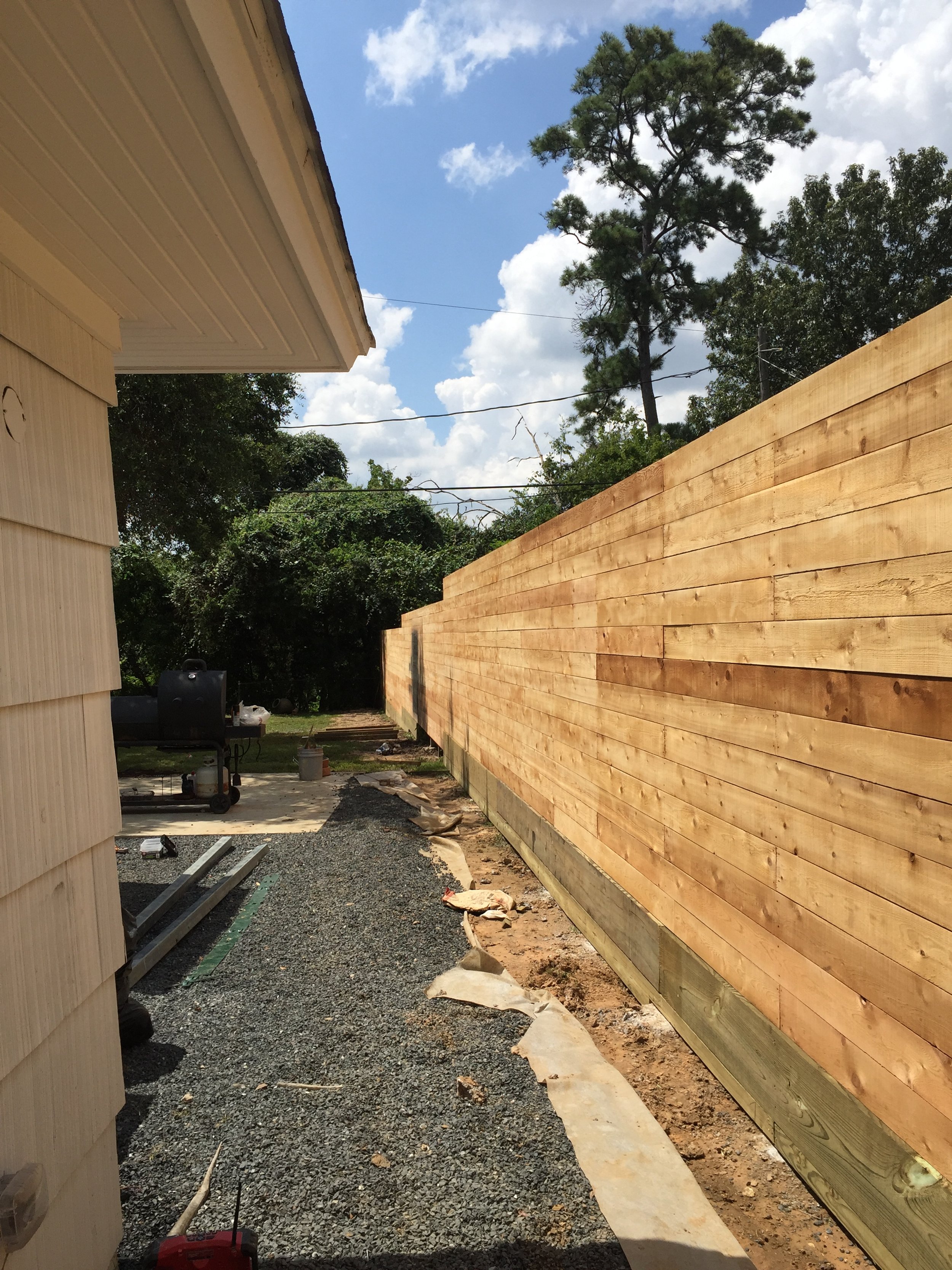 Wooden fence along a gravel path next to a house under a blue sky with clouds.