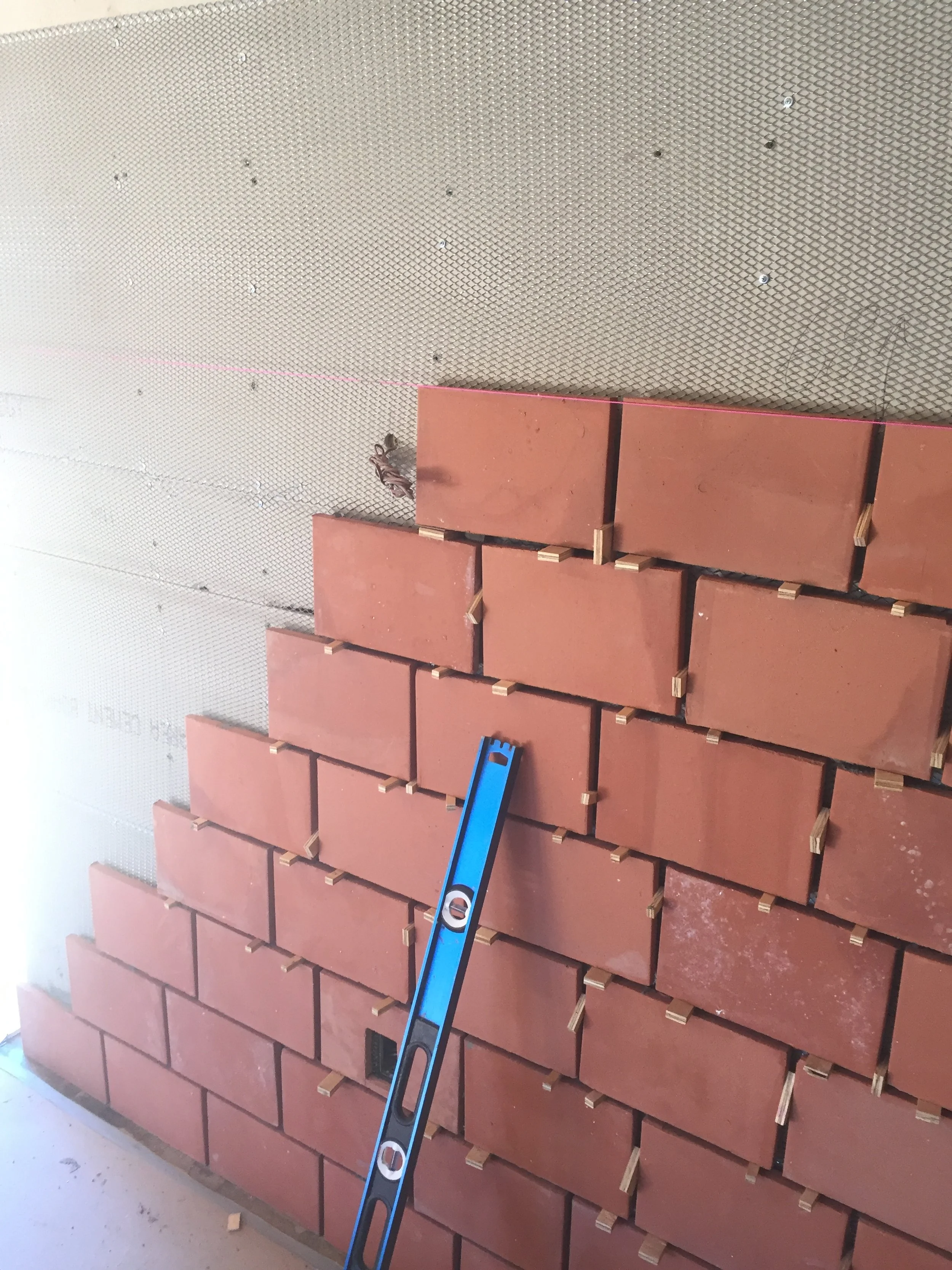 Partially constructed brick wall with a blue level leaning against it and a metal mesh backing. Wooden spacers are visible between bricks.