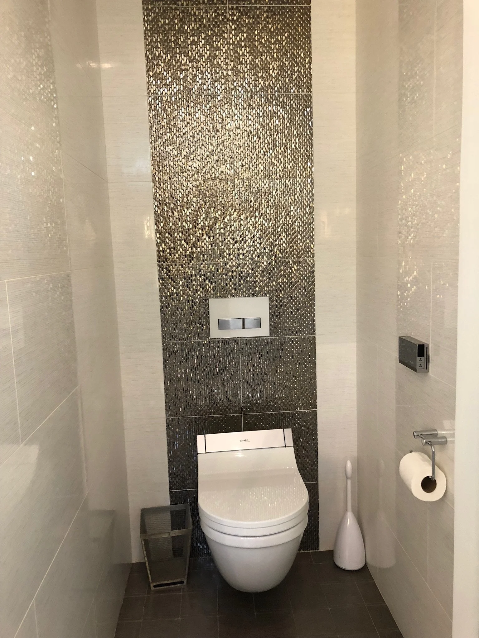 Modern bathroom with wall-mounted toilet, mosaic tile backsplash, toilet paper holder, and toilet brush.
