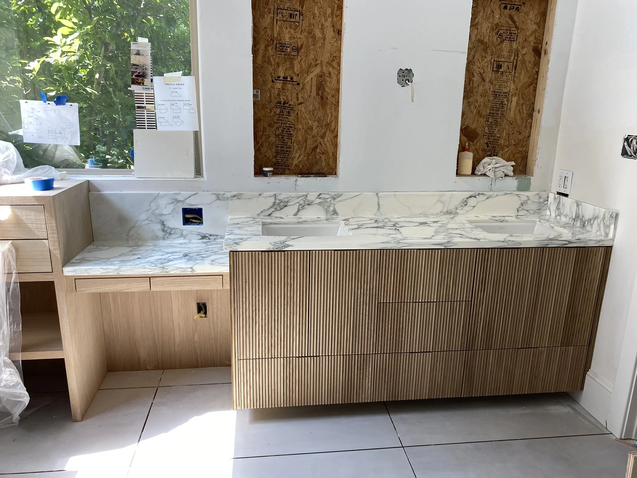Bathroom under renovation with marble countertop, wooden cabinets, and white tile floor.