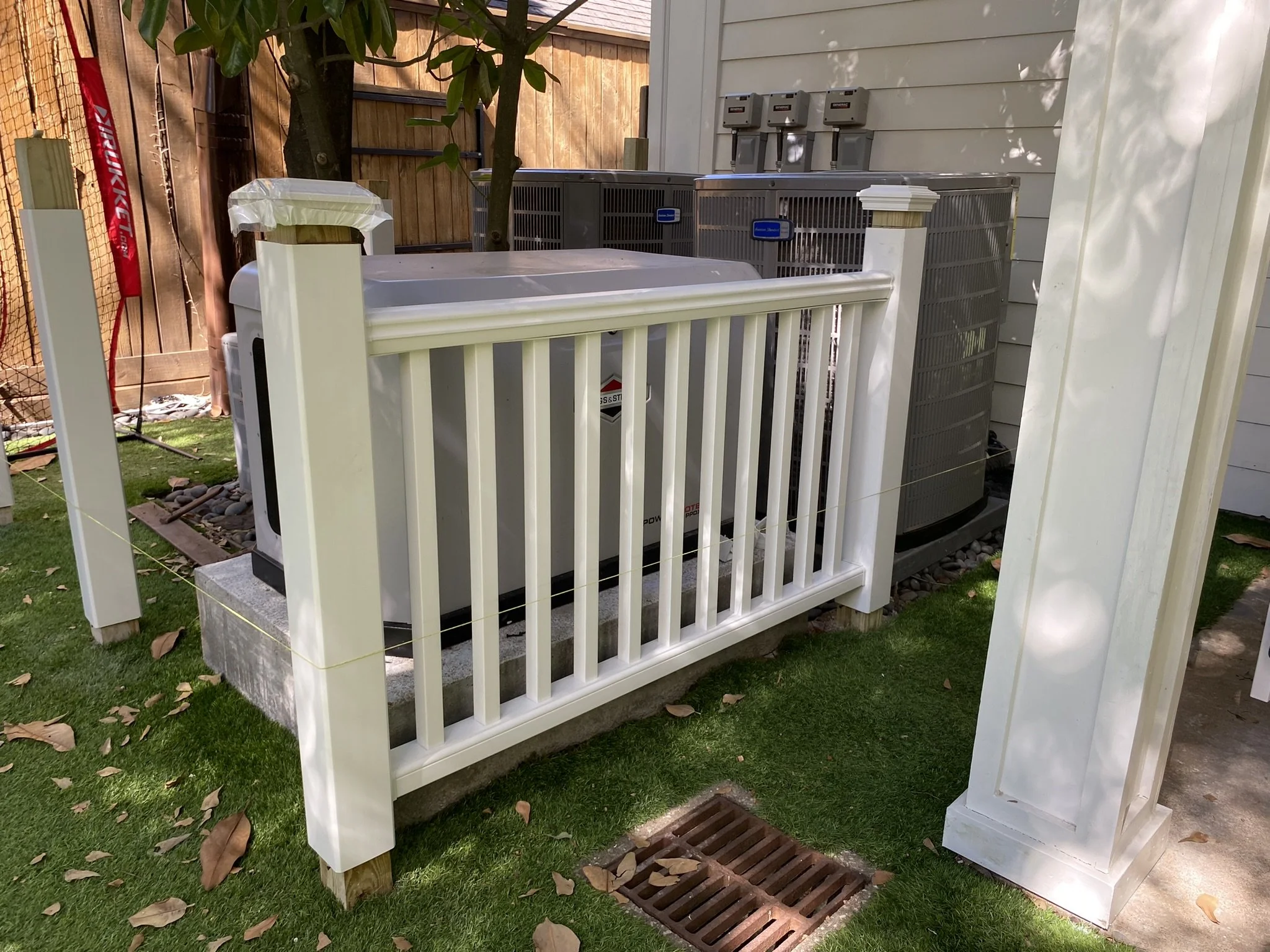 Outdoor air conditioning units and generator behind a white fence on artificial grass, next to a wooden shed.