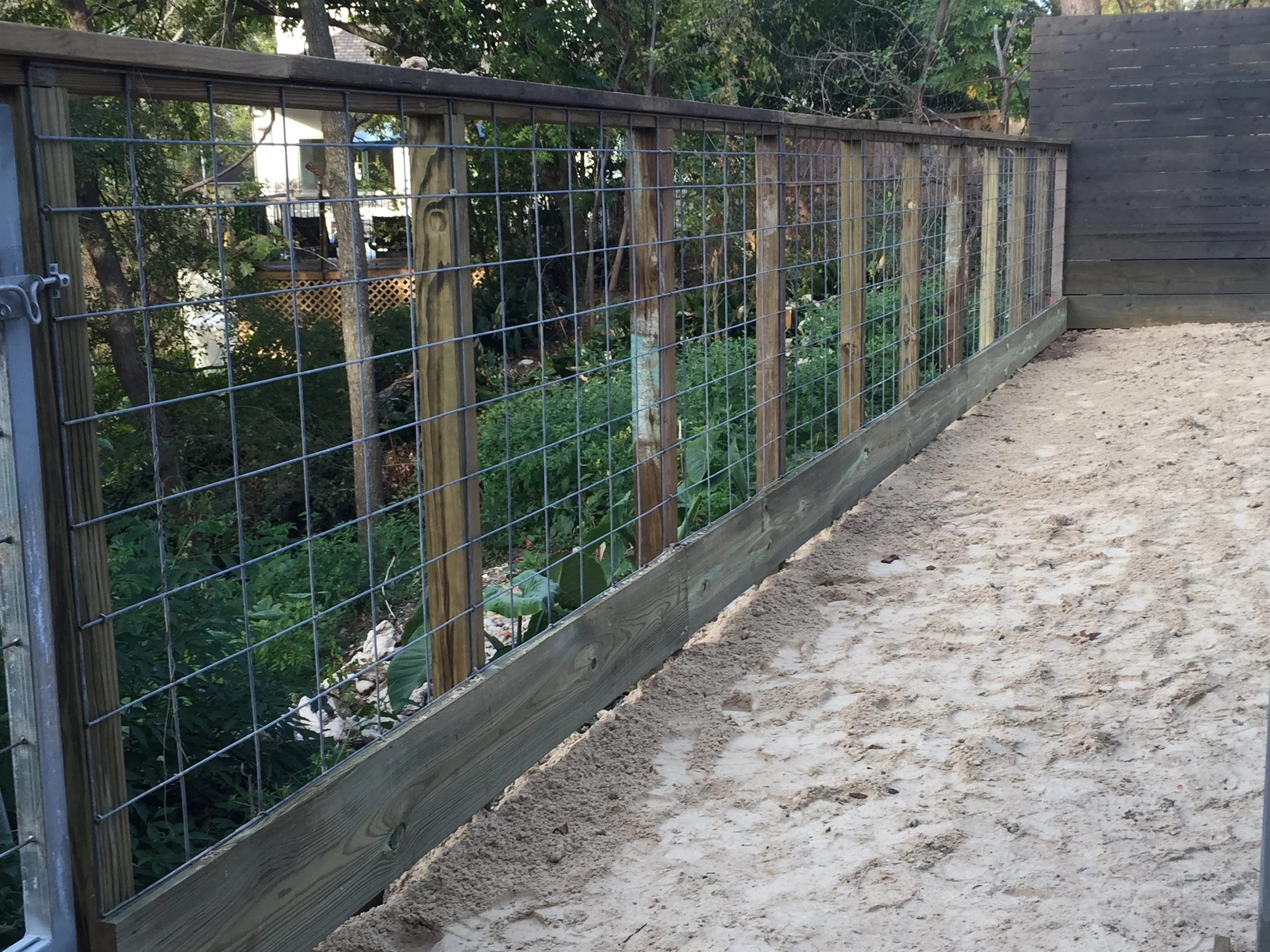 A wooden and wire mesh fence set on sandy ground, with trees and greenery in the background.