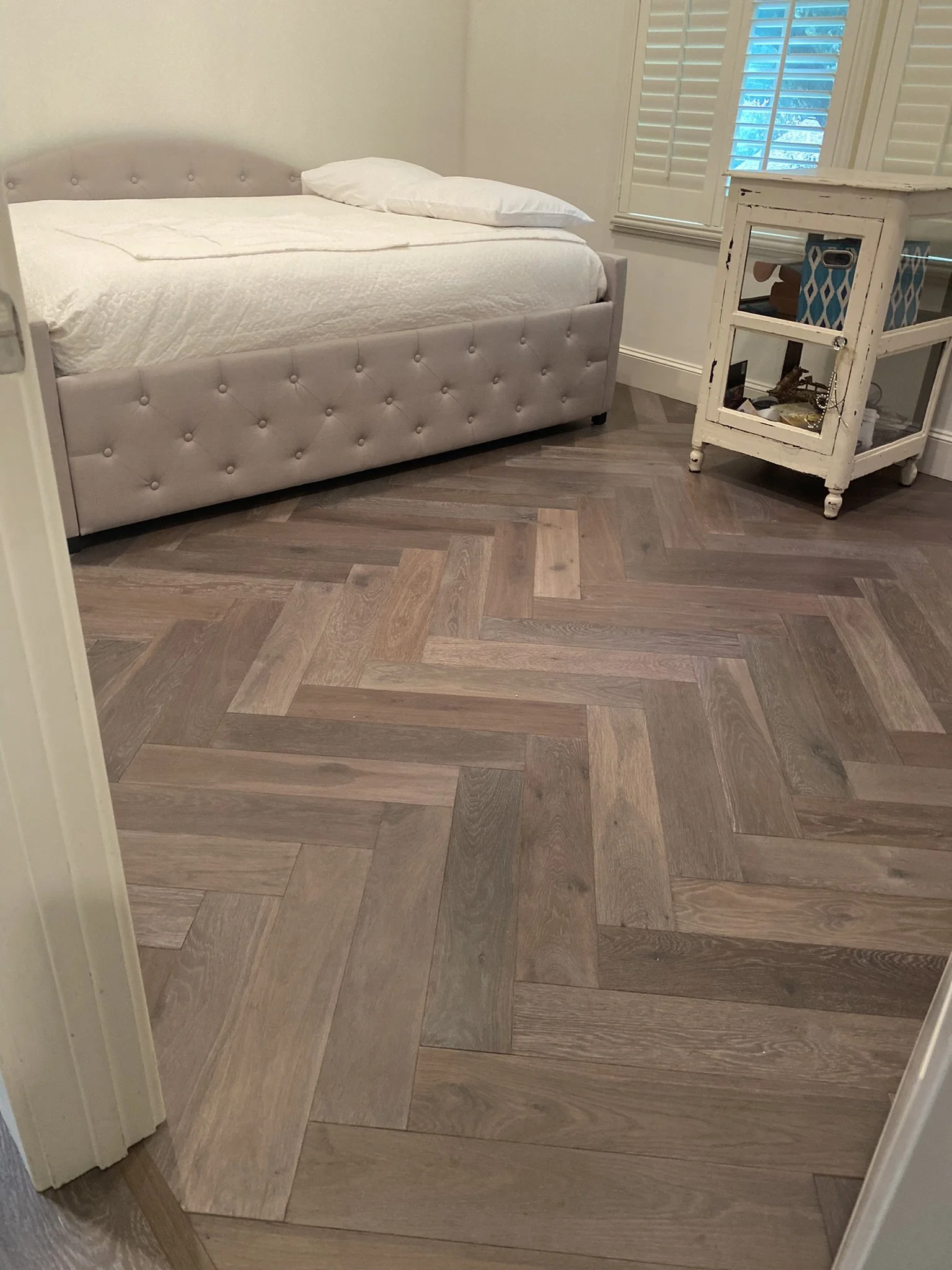 Bedroom with herringbone wood floor, tufted bed, and small white cabinet.