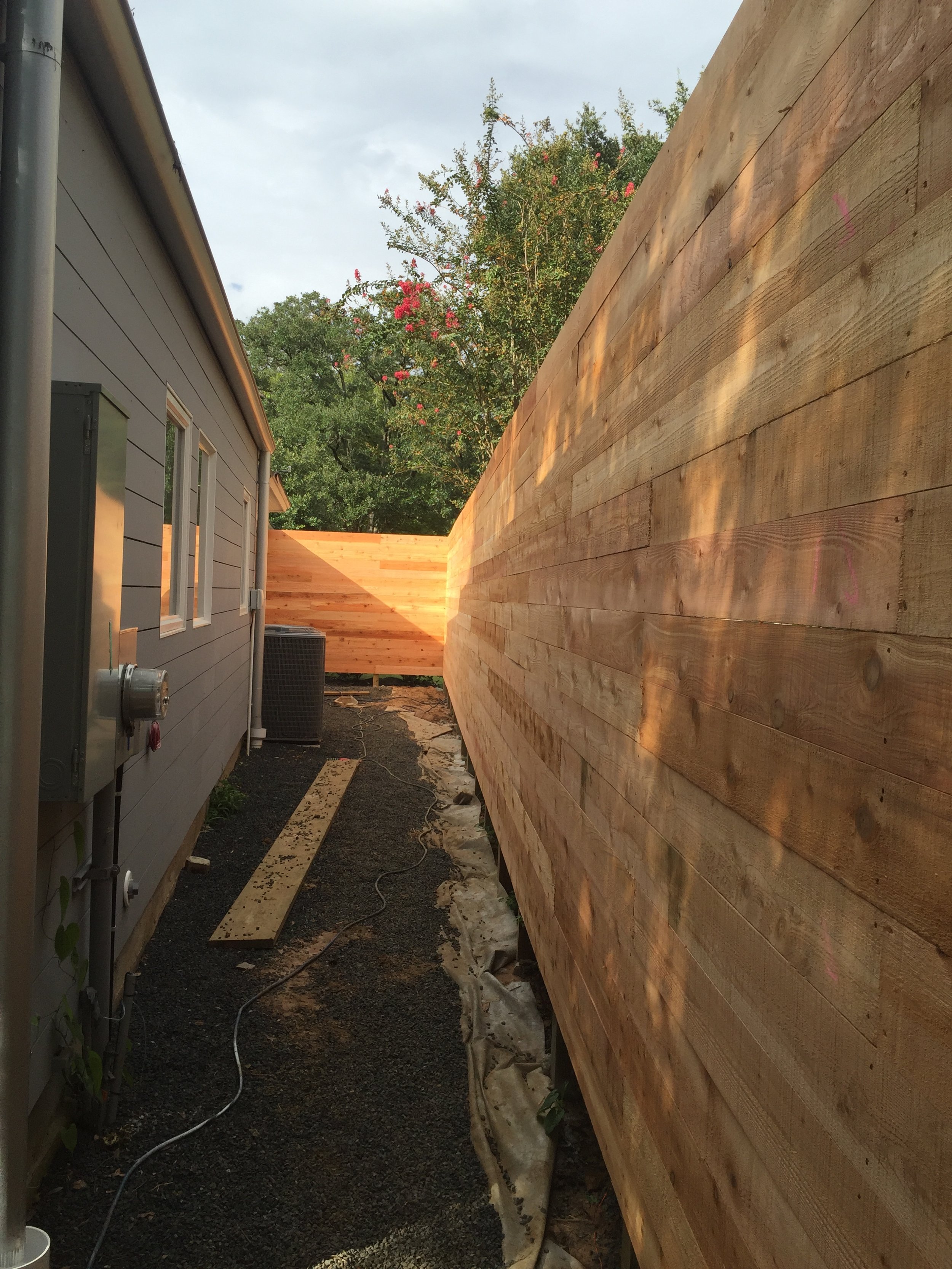Narrow passage between a building and a wooden fence, with gravel path and greenery in the background.