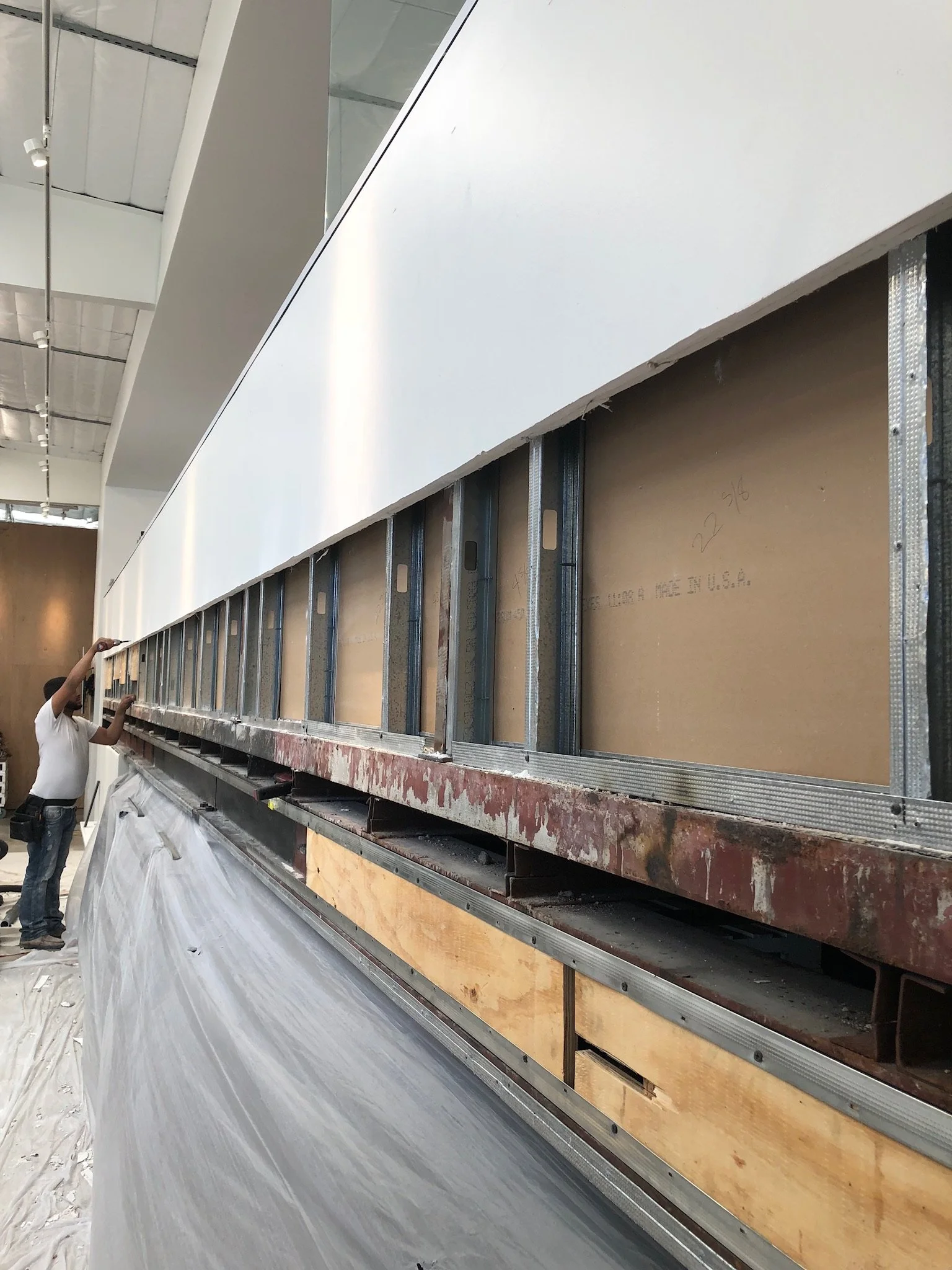 Construction worker adjusting metal framing and drywall in a large interior space with exposed ceilings and lighting.