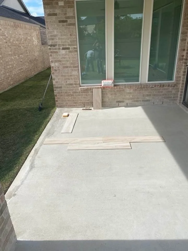 Outdoor patio area with unfinished tile flooring next to a brick wall, featuring large windows. Some tiles are placed on the concrete surface, and a broom is seen leaning against the wall. Sunlight is casting shadows on the ground.