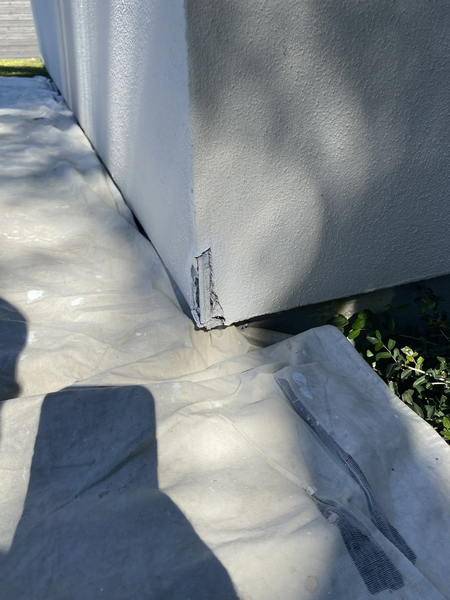 Corner of a white stucco wall with a small damaged area at the base, placed on a ground covered with protective tarp, with plants on one side.