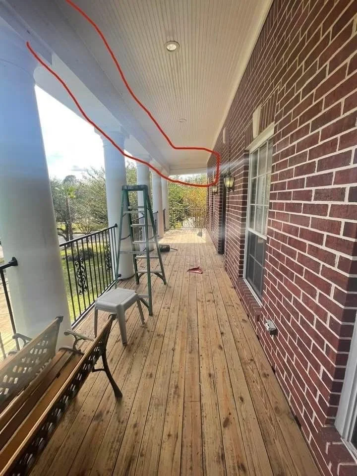 A wooden porch with a brick wall, white pillars, and a ceiling. A ladder and a stool are placed on the porch, and there's a red line drawn on the ceiling. A bench and railing are visible on the left side.