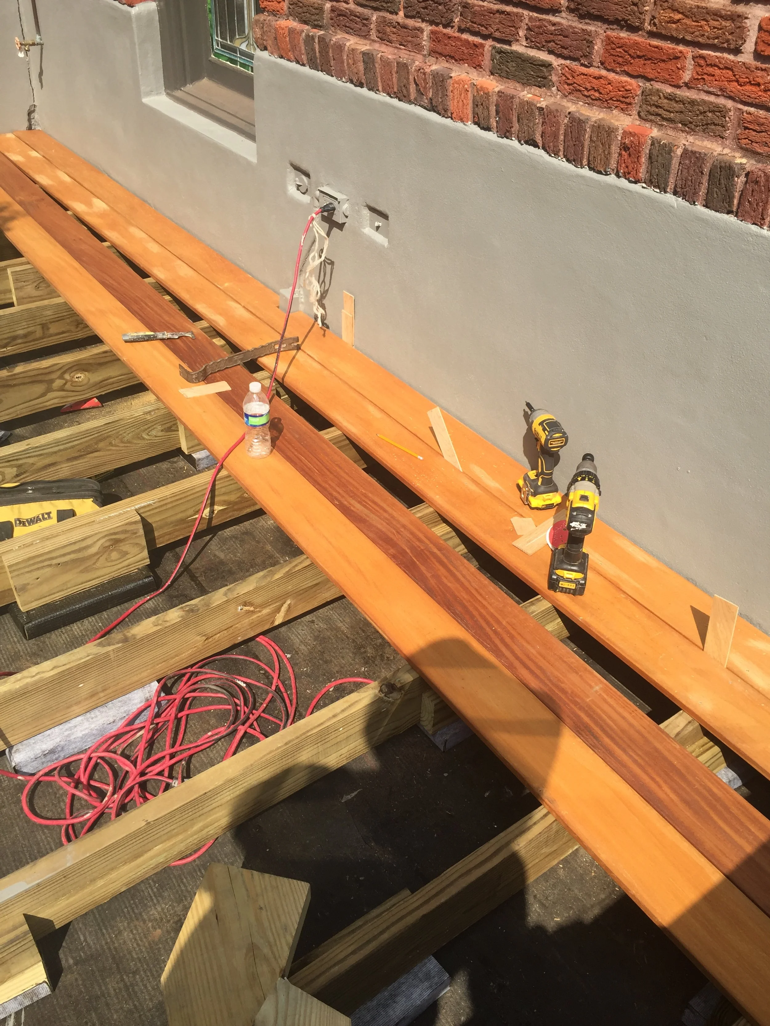Wooden deck construction with tools, red extension cord, and partially built decking boards against a brick wall.