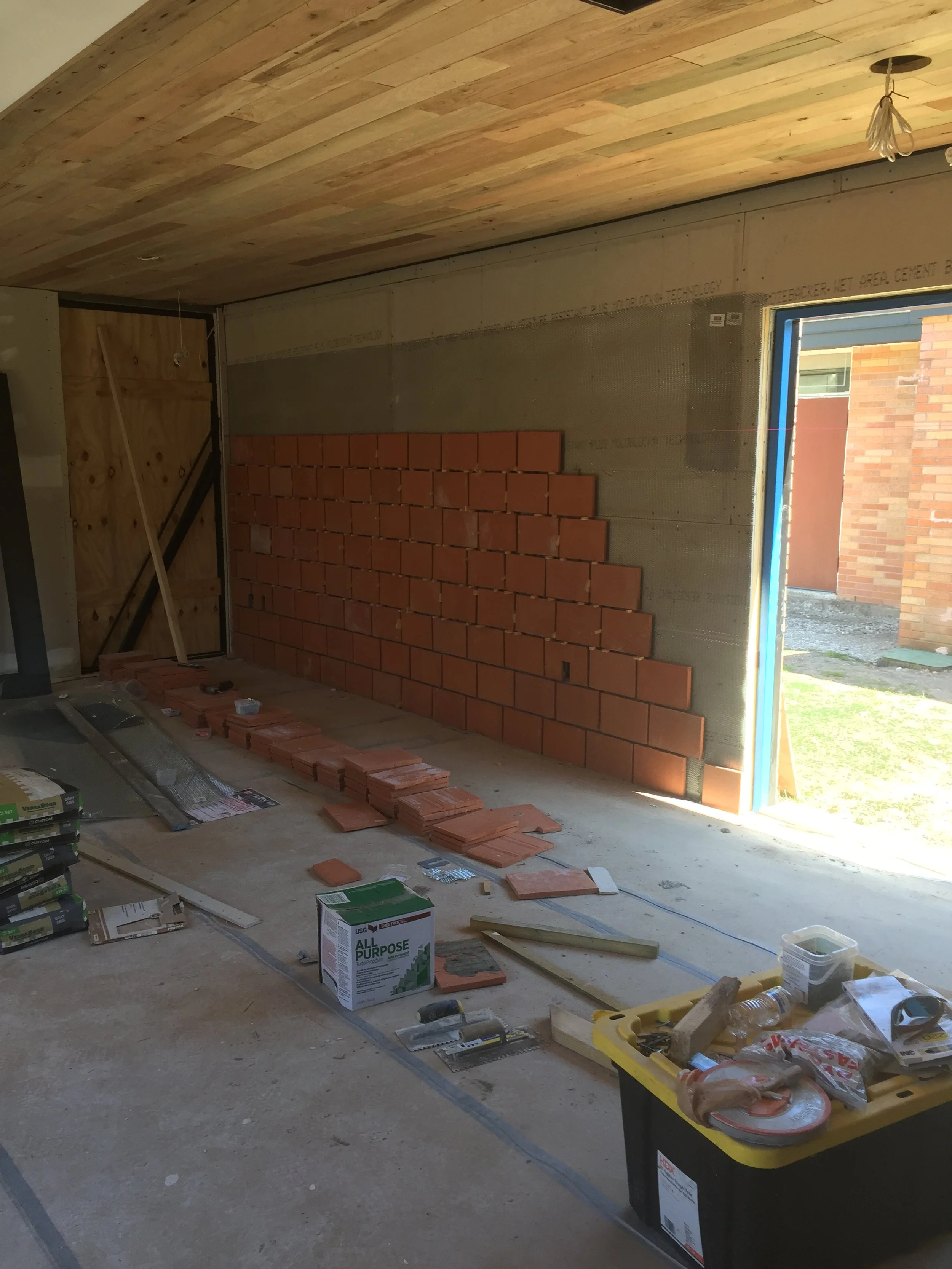 Interior construction site with brick wall in progress, wooden ceiling, building materials, tools, and a partially open doorway.
