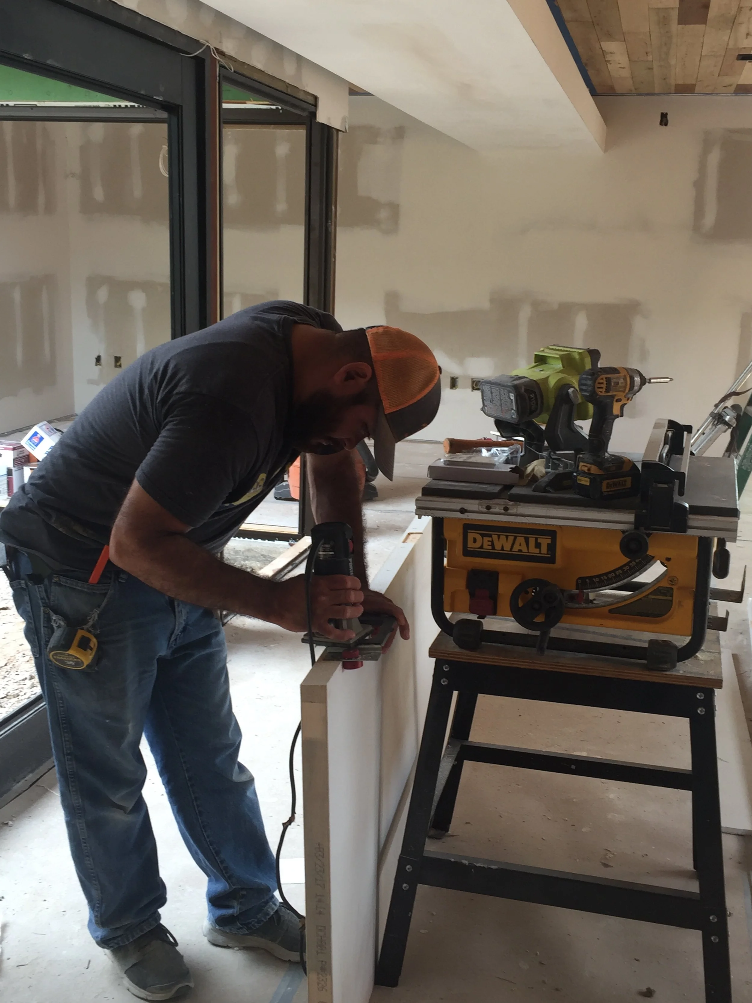 A man using a power tool on a wooden board in a workshop with a DeWalt table saw nearby.